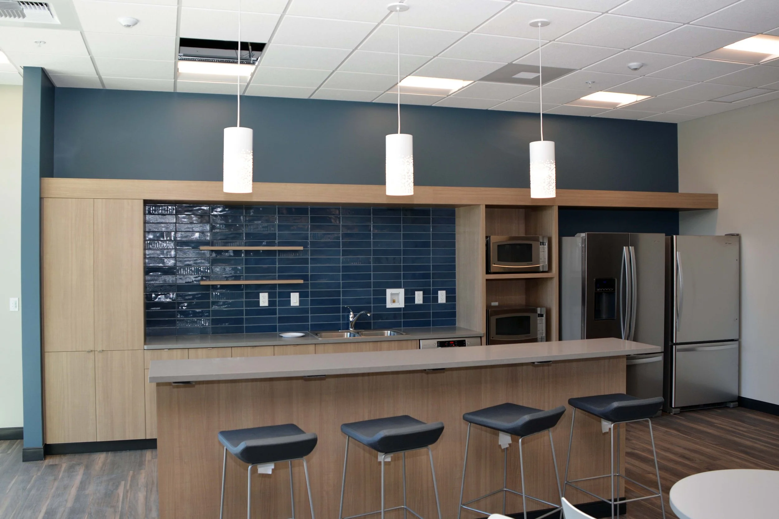 Modern kitchen with wooden cabinets, blue tiled backsplash, stainless steel appliances including a microwave and refrigerator, white pendant lights, a countertop, and barstool seating.