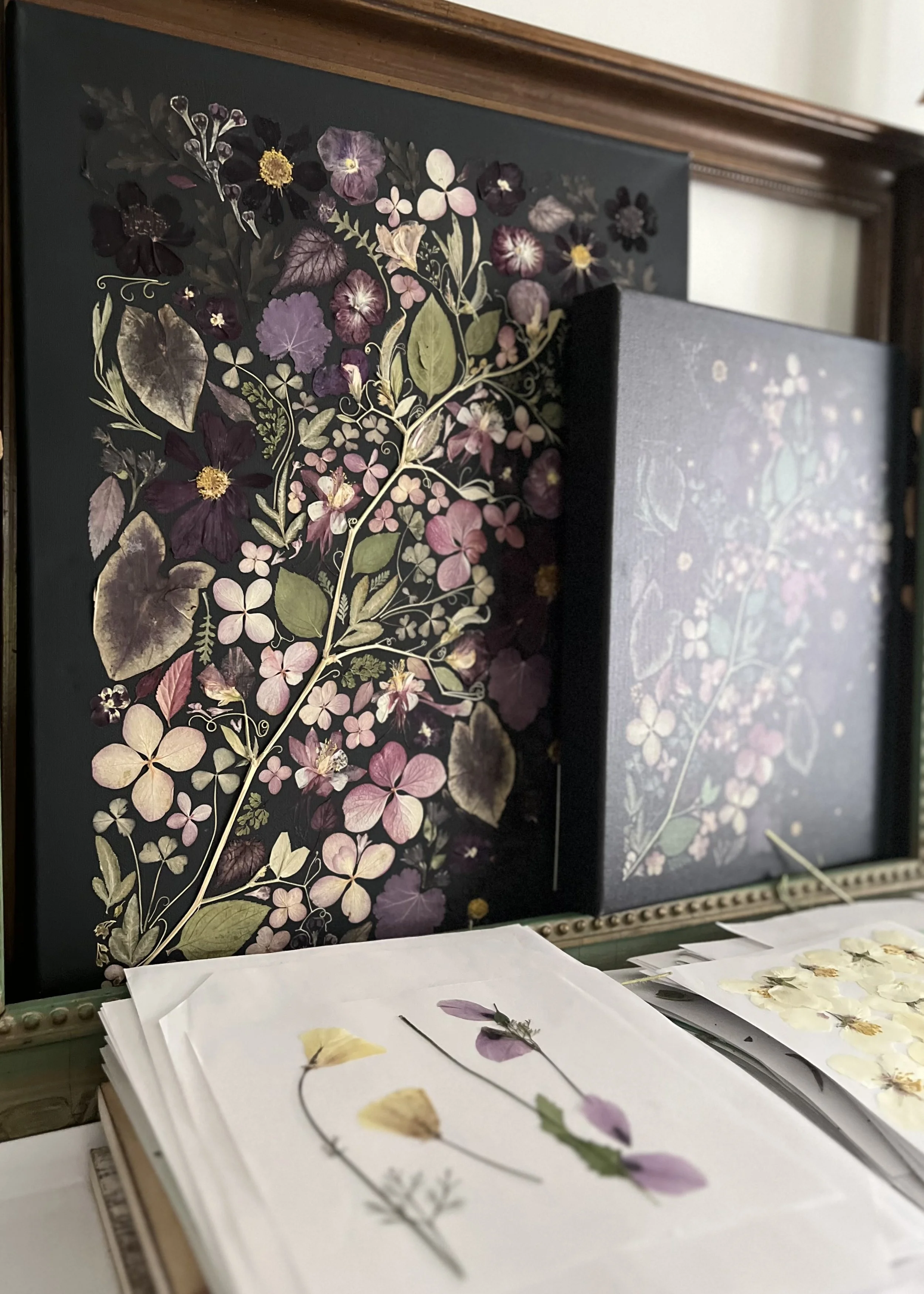 a black and moody piece of art with a second  piece of art of the same and pressed flowers at the base of the photo.