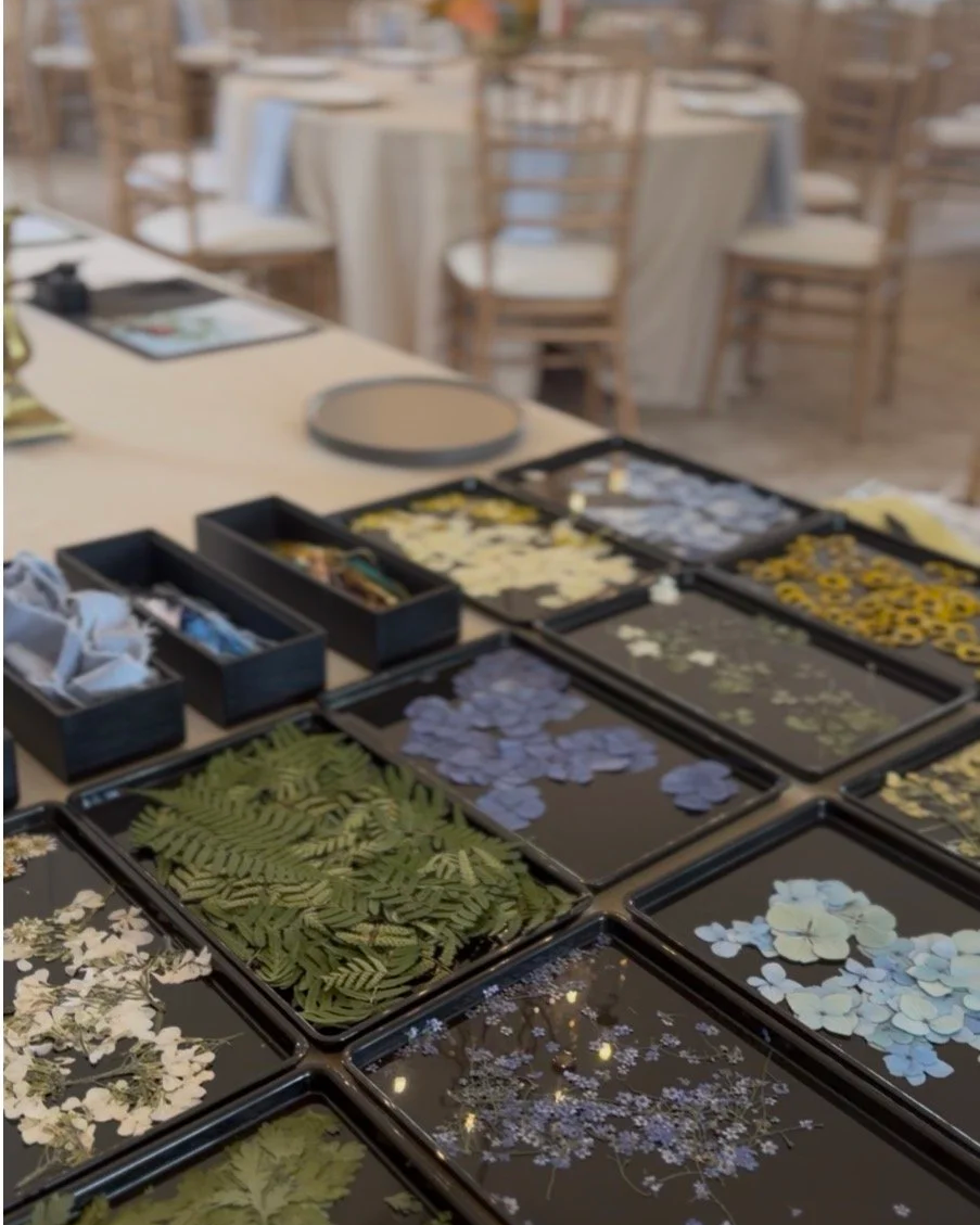 Table with various pressed flowers and greenery organized in black trays, set up in a bright, elegant event space with wooden chairs and tables in the background.
