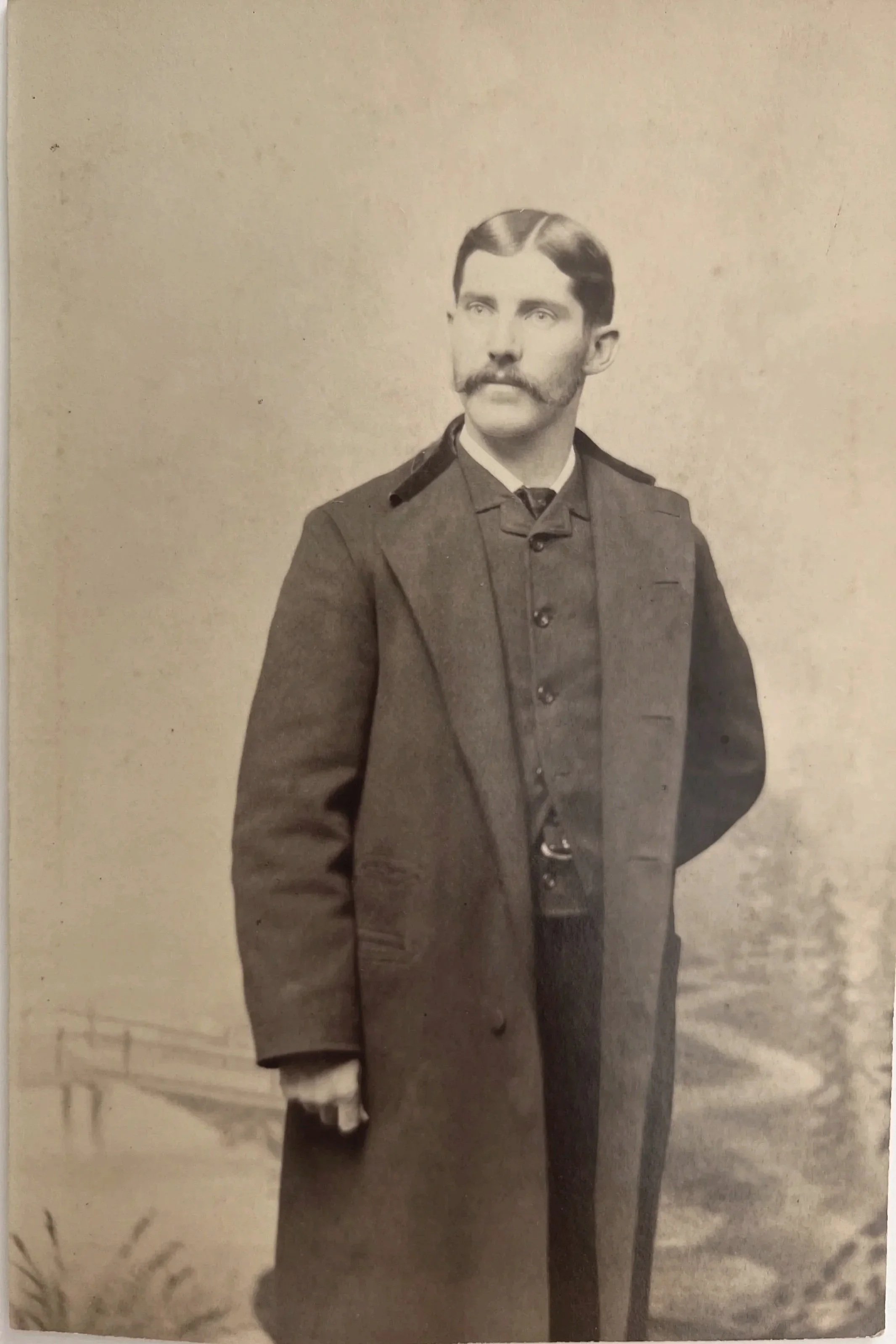 A black-and-white portrait of a man dressed in a military uniform with a long coat, vest, and tie, standing with one hand behind his back, against a light background with faint outdoor scenery.