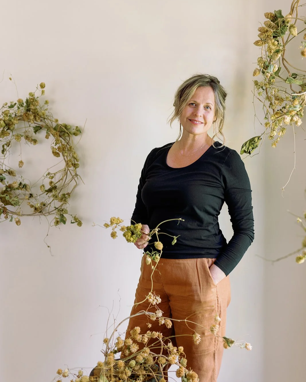 A woman with blonde hair wearing a black long-sleeve shirt and brown pants standing in front of a plain light-colored wall, surrounded by dried plants and flowers.