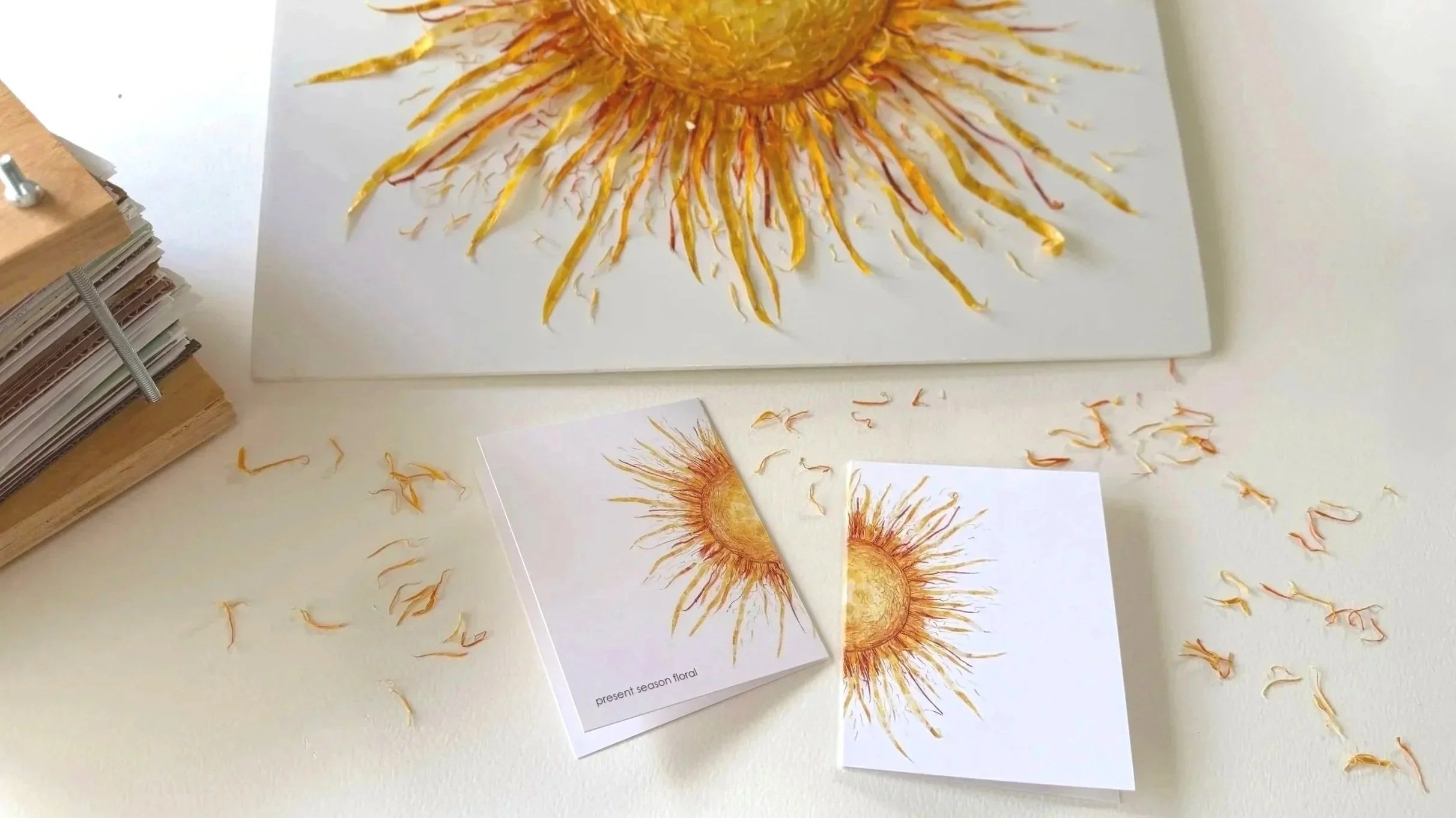 A drawing of a sunflower with yellow petals and a bright sun-like middle on a white piece of paper, surrounded by sunflower petal pieces and other flower pieces.