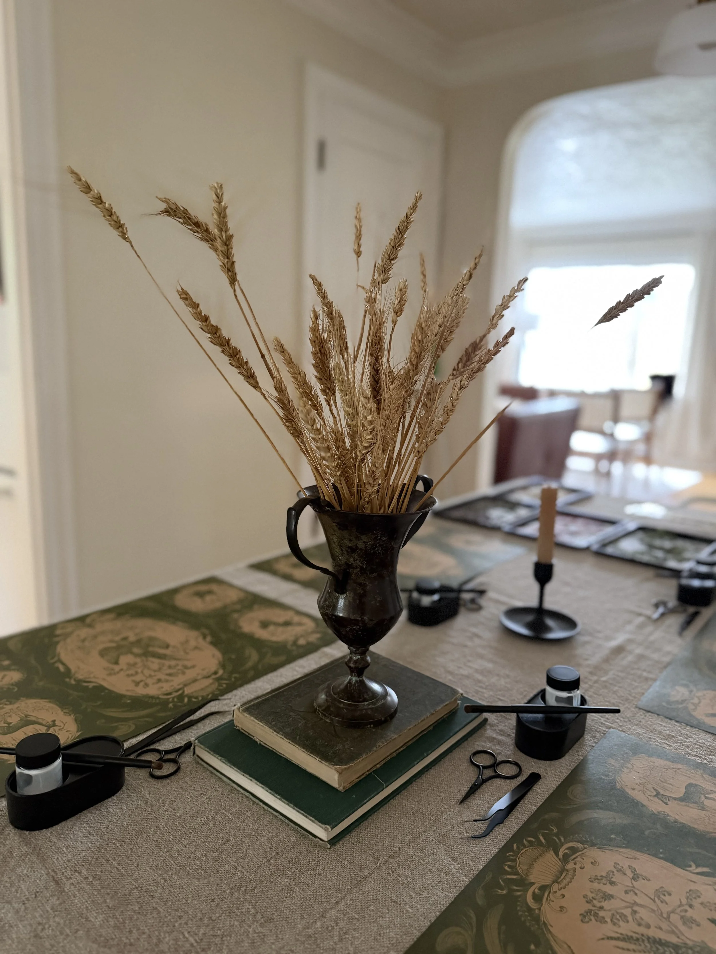 A table with various art and craft supplies, including a black vase filled with dried wheat stalks, books, scissors, an artists' palette, paint tubes, and printed images, set in a room with a window and chairs in the background.