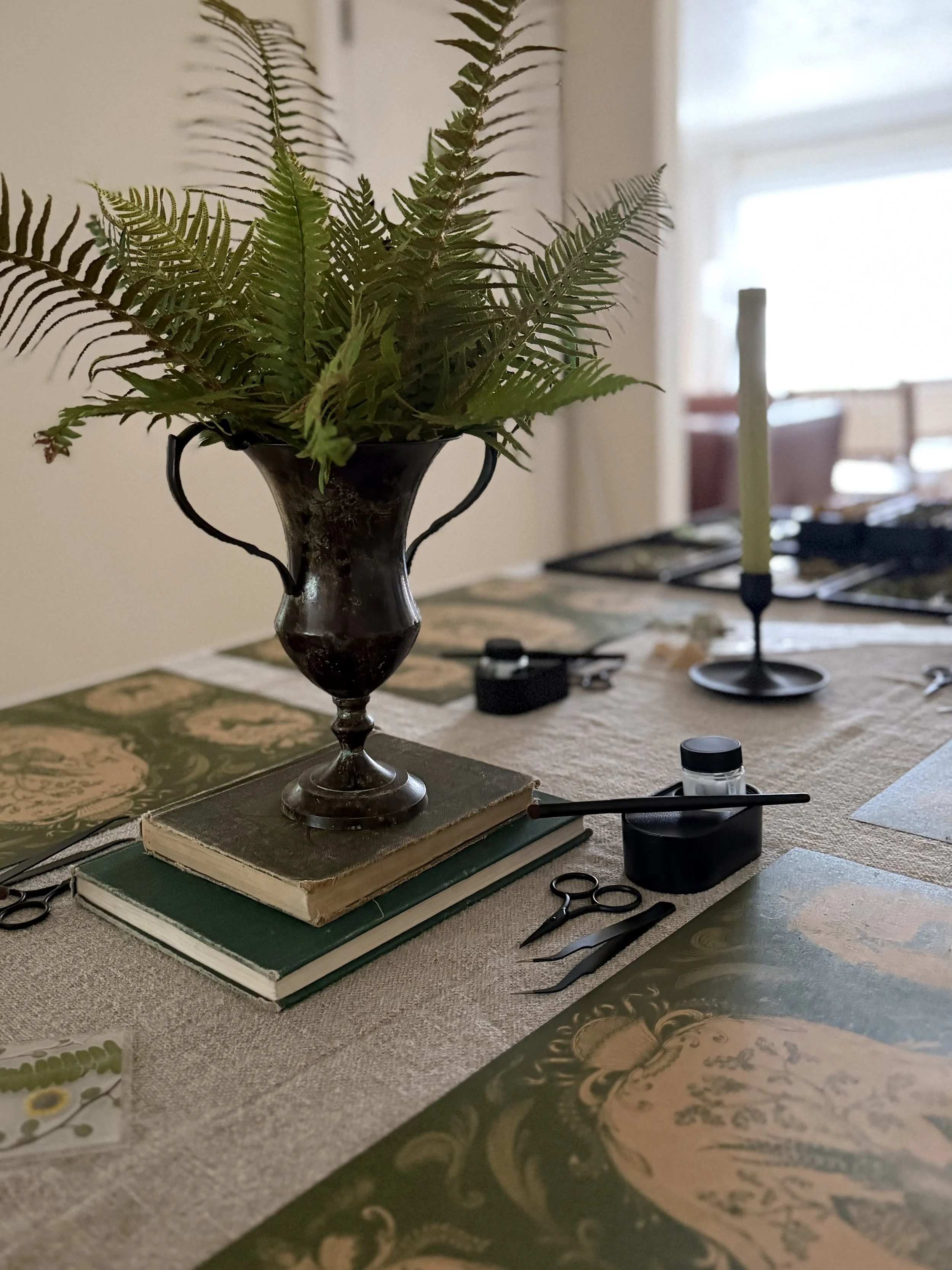 A table with a vintage vase holding fern leaves on top of a stack of two old books. The table contains scissors, a small black container, and other stationery. In the background, there is a tall candlestick with a candle, and a window with blinds.