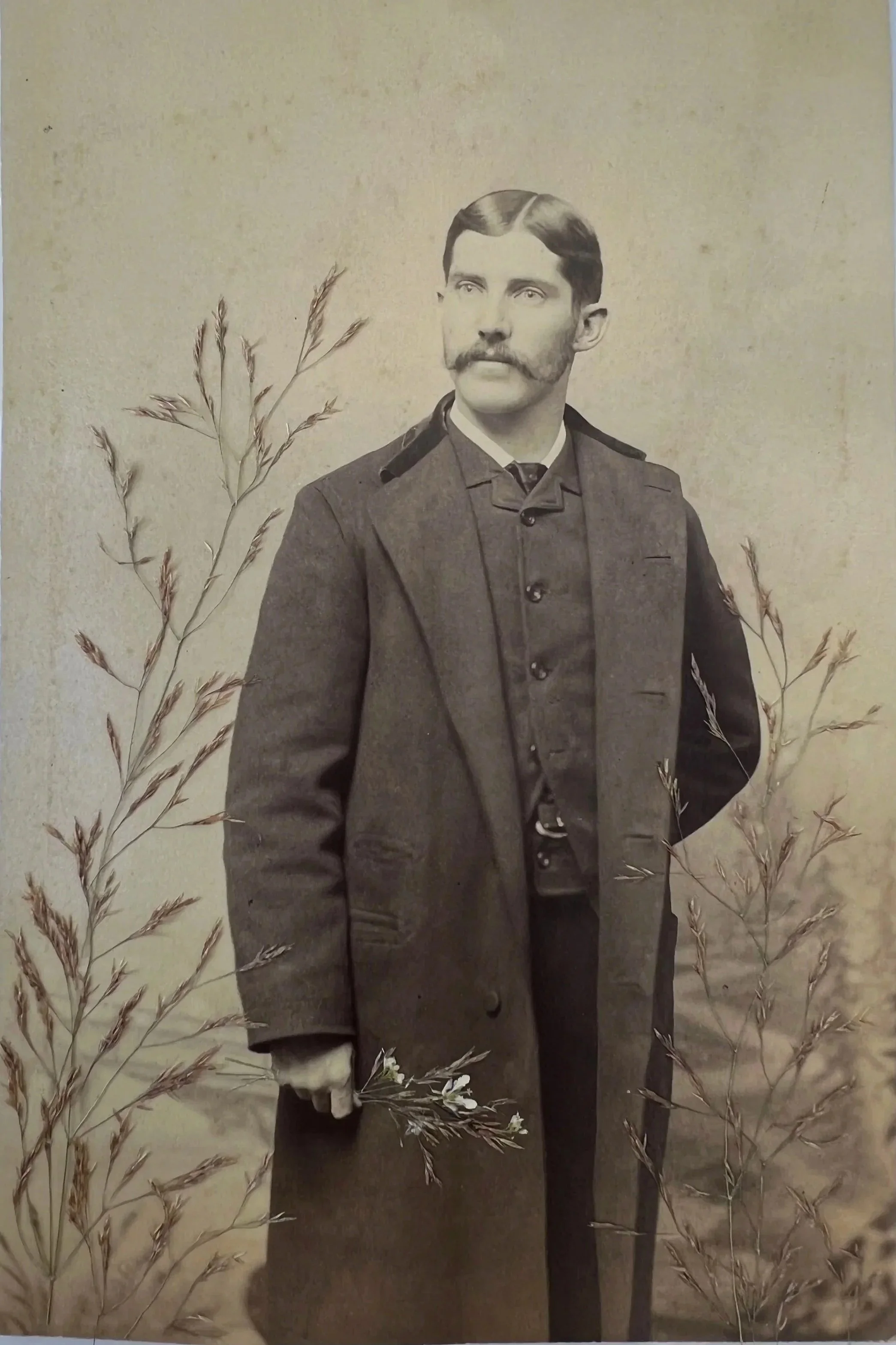 A black and white vintage portrait of a man in a military uniform with a long coat, holding a flower in his right hand, standing among tall grass.