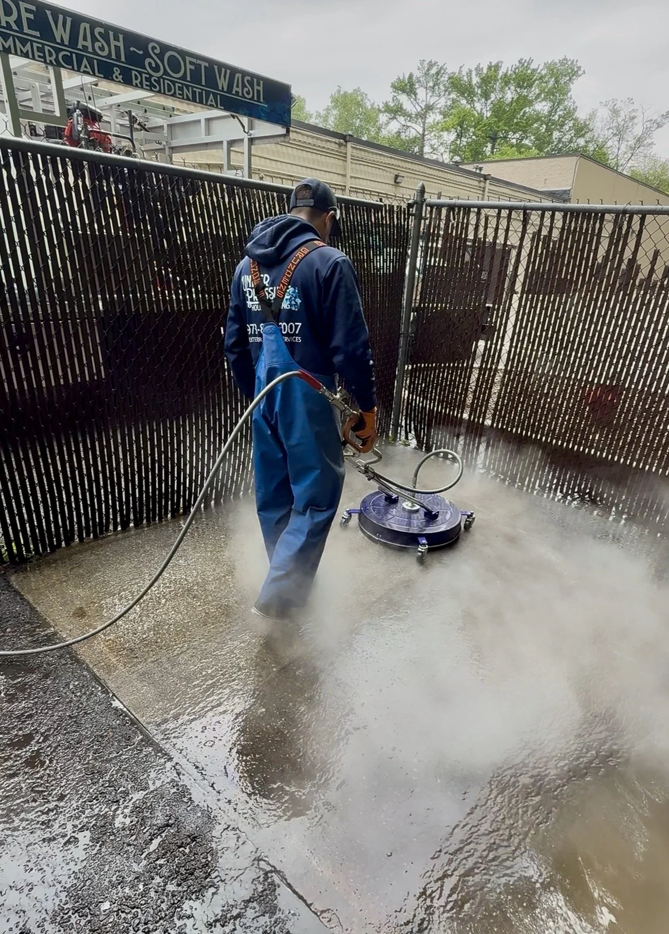A person wearing a dark hoodie, blue coveralls, and gloves is power washing a concrete outdoor surface, creating a mist and cleaning the area near a black metal fence.