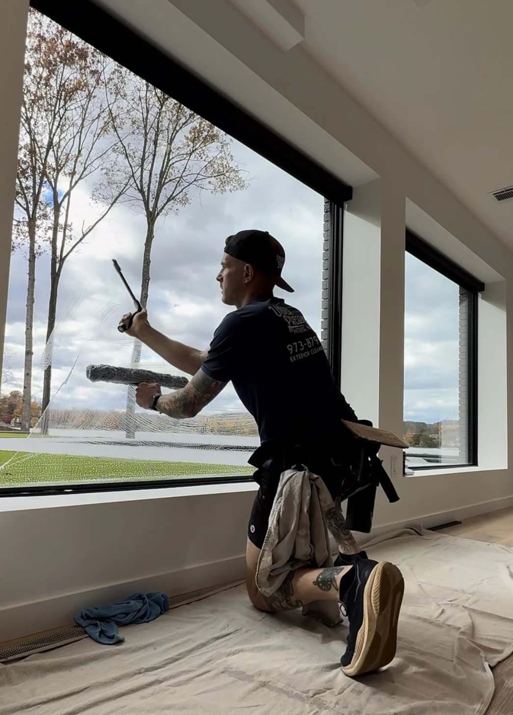 A man kneeling on a floor with a cloth over it, painting large window panes from inside a modern building, with an outdoor scene of trees and a cloudy sky visible through the windows.