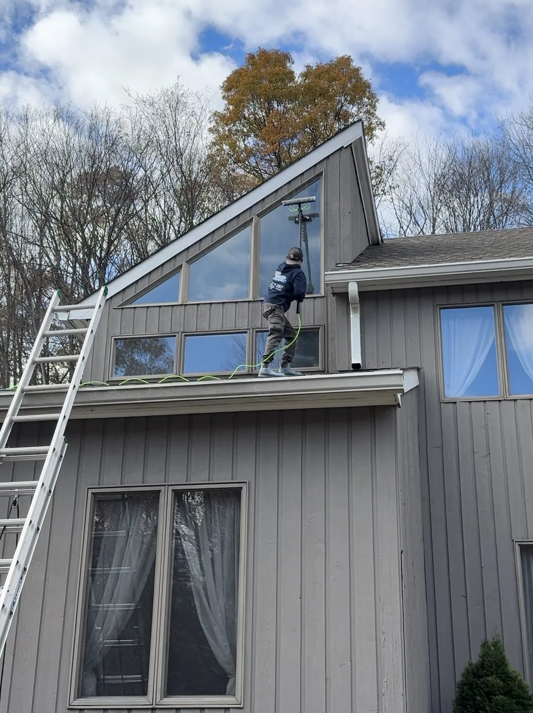 A person cleaning windows on a two-story house with a pole and spray device, standing on the roof edge, with a ladder leaning against the house, trees, and a partly cloudy sky in the background.