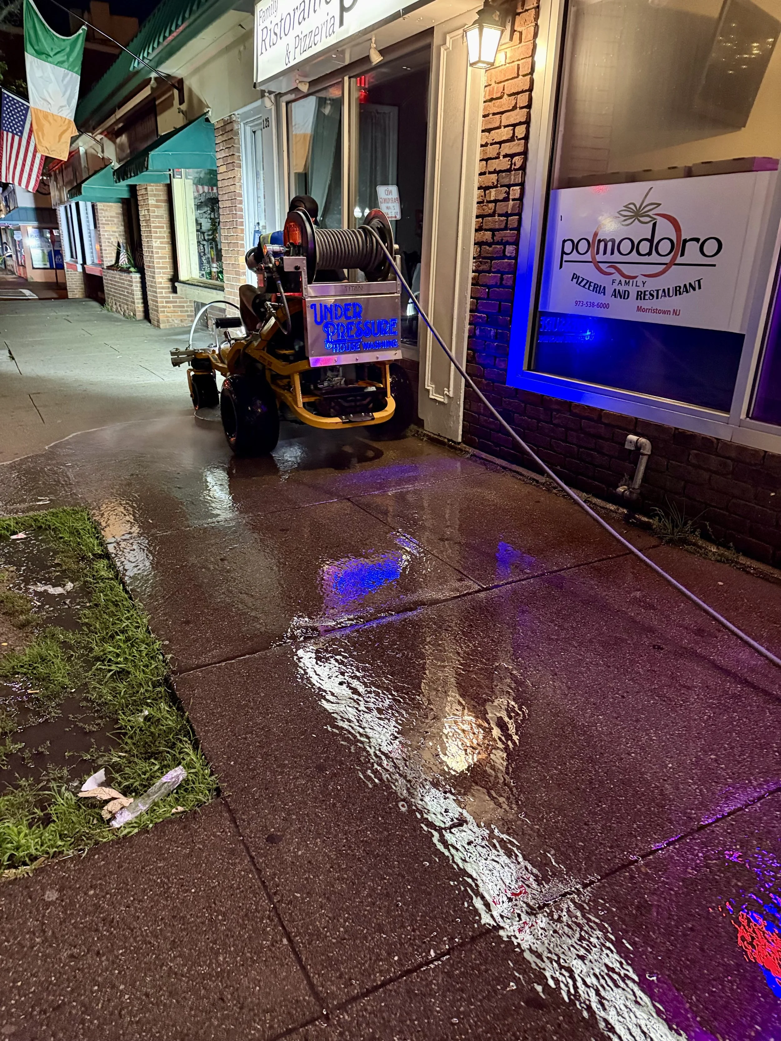 A street sidewalk at night with a water wet surface reflecting colorful neon lights from nearby signs. There is a pizza restaurant called 'Pomodoro' with a lit sign, and a large cleaning machine with a hose attached is parked outside, with a sign on it indicating 'Under Pressure House Washing.' The building has brick walls, and the street features small flags hanging above.