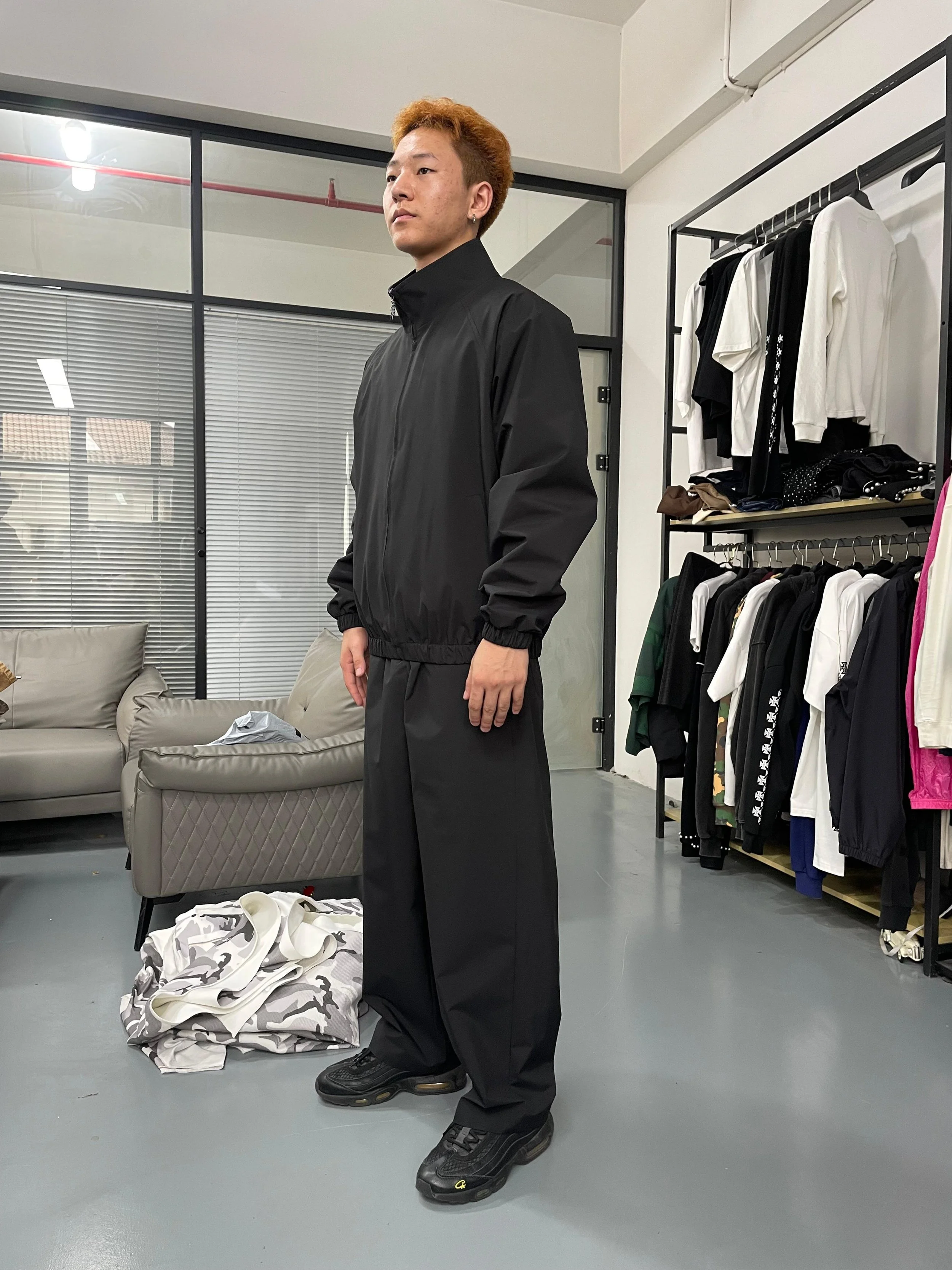 A young man with reddish-brown hair, wearing a black windbreaker jacket and black pants, standing indoors in a clothing store or dressing room area.