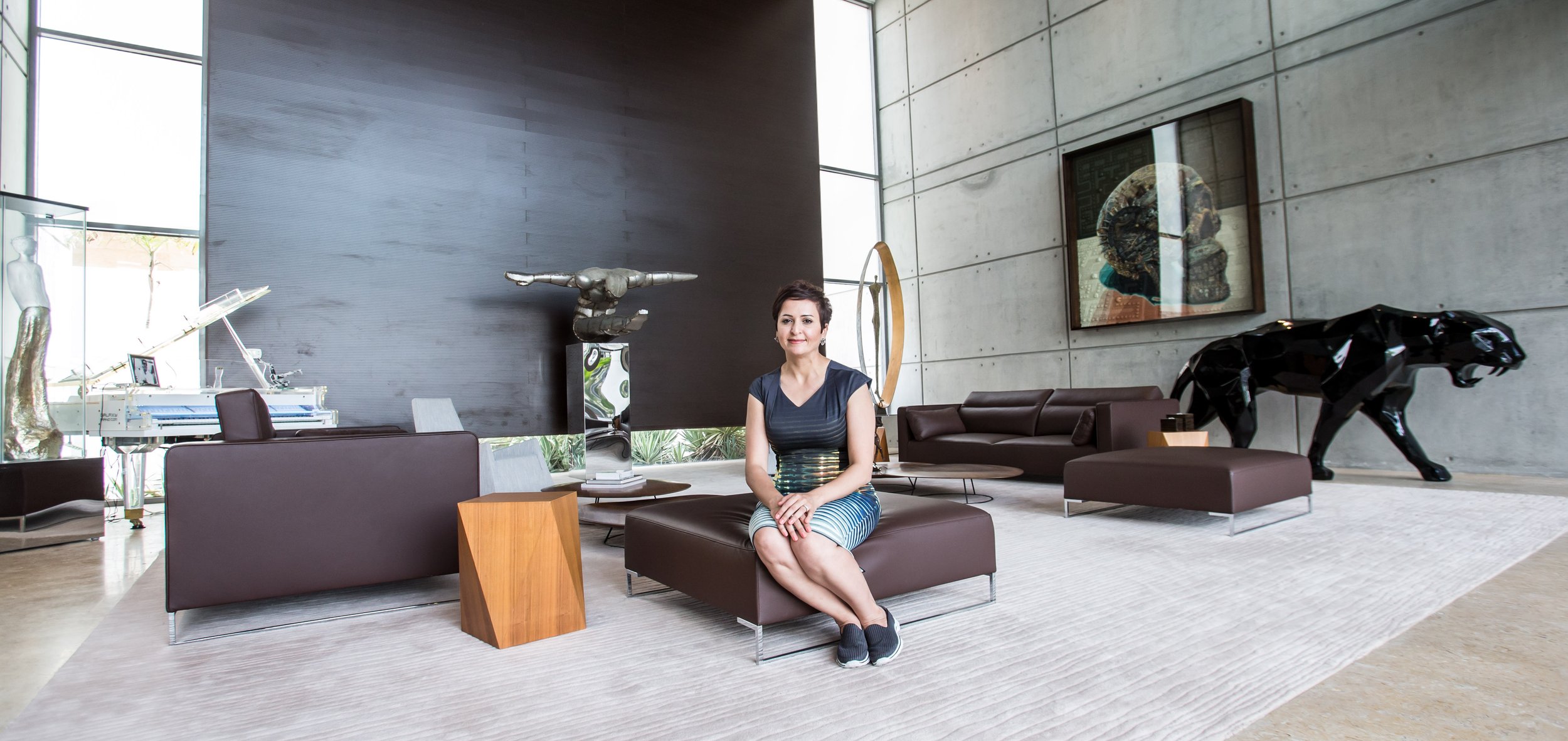 A modern living room with a woman sitting on a brown leather ottoman. The room features contemporary furniture, a white grand piano, a large black sculpture of a panther, and various abstract artworks on a concrete wall.