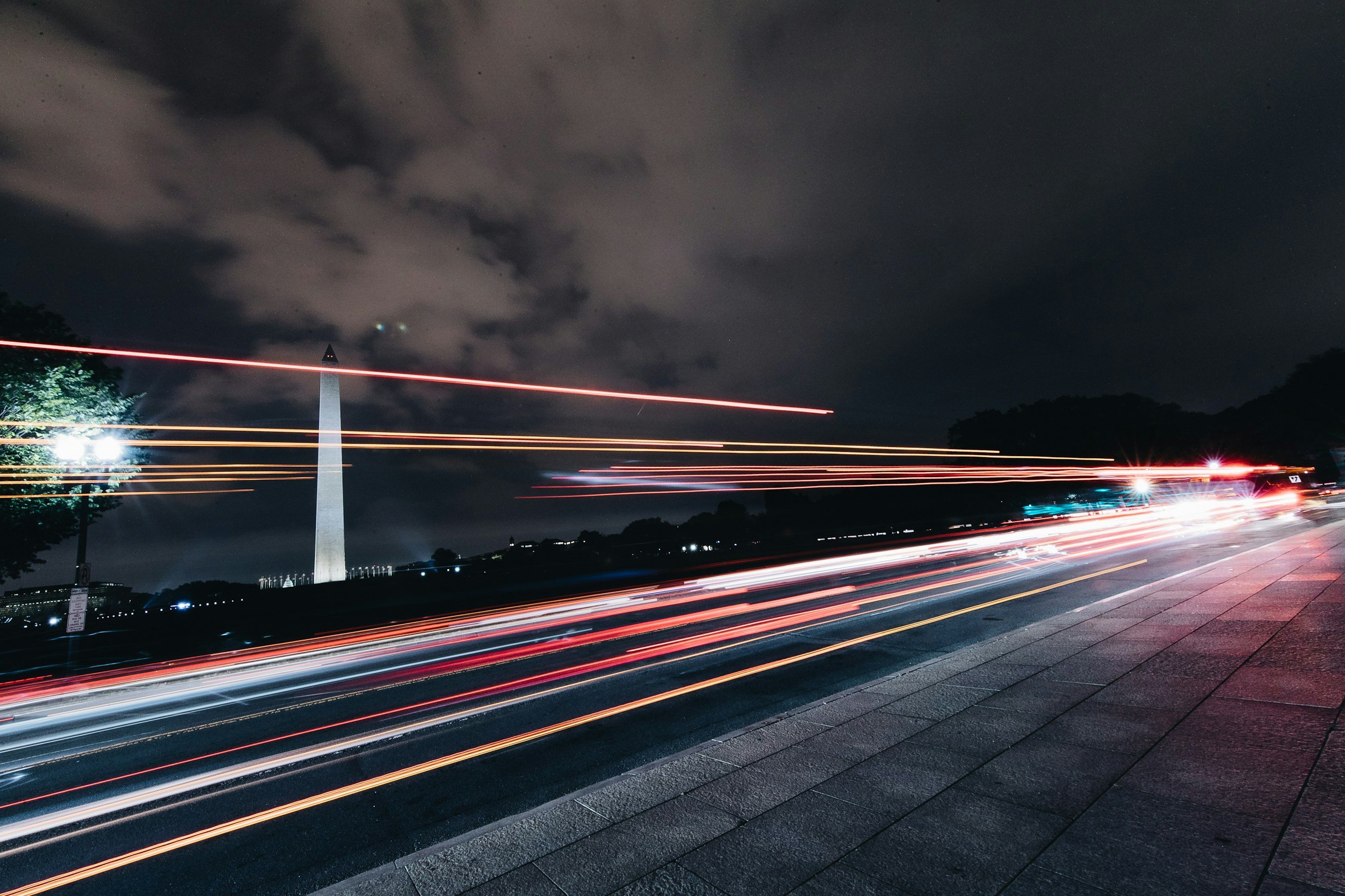 Nighttime city street with light trails from moving vehicles, Washington Monument in the background, cloudy sky, and sidewalk on the right.