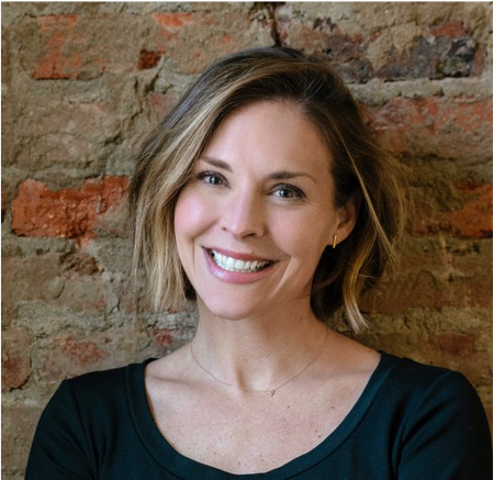 A woman with shoulder-length light brown hair smiling in front of a brick wall, wearing a black top.