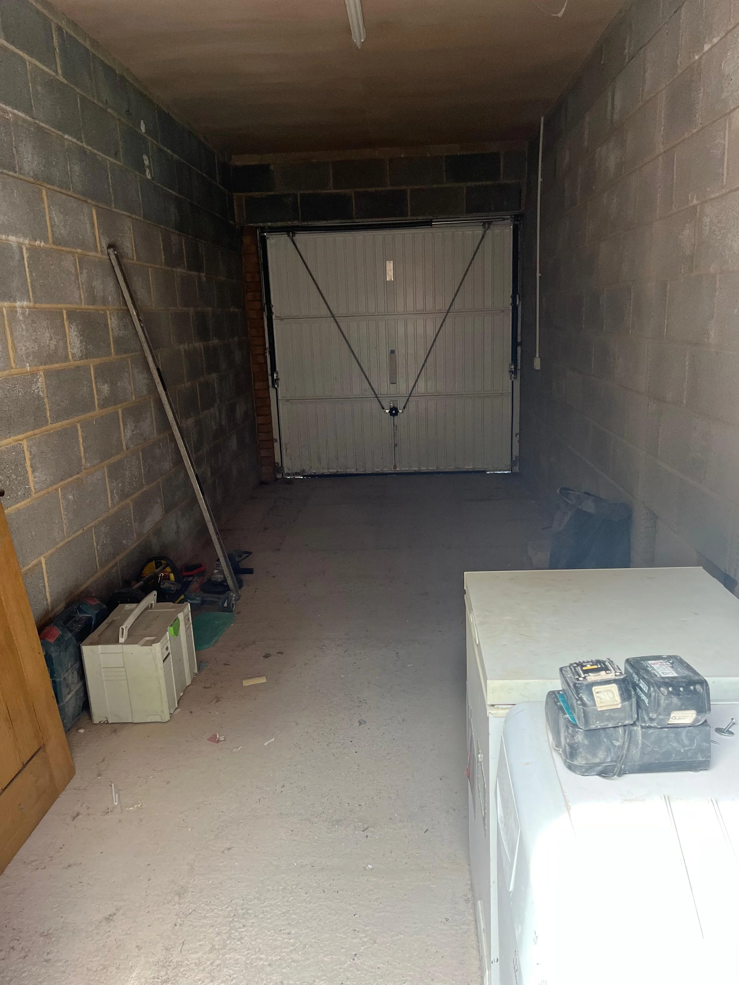 An unfinished garage with concrete block walls, a white garage door, a white table with tools, and various tools and equipment on the floor and table.