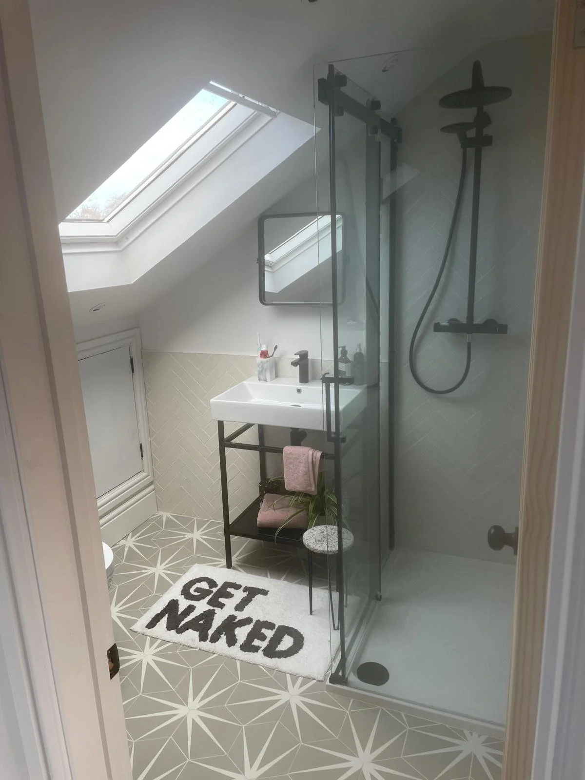 A modern bathroom with skylight, glass shower enclosure, and a small white sink. There are decorative tiles on the floor and wall, a black towel rack, and a bath mat with the words 'GET NAKED'.
