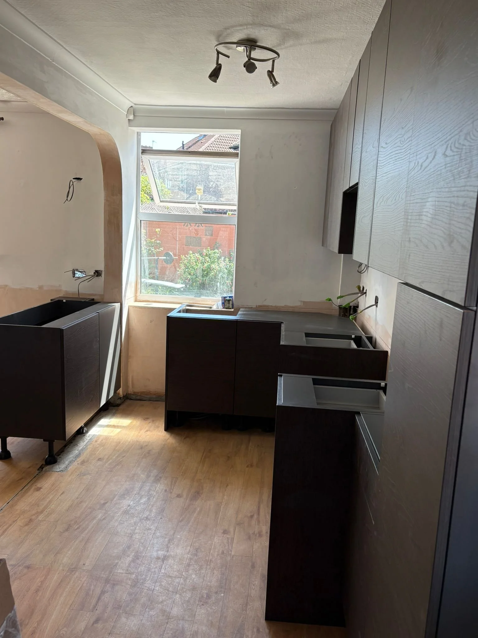 Empty kitchen with dark cabinets, a large window, and wooden flooring, with some construction or renovation in progress.