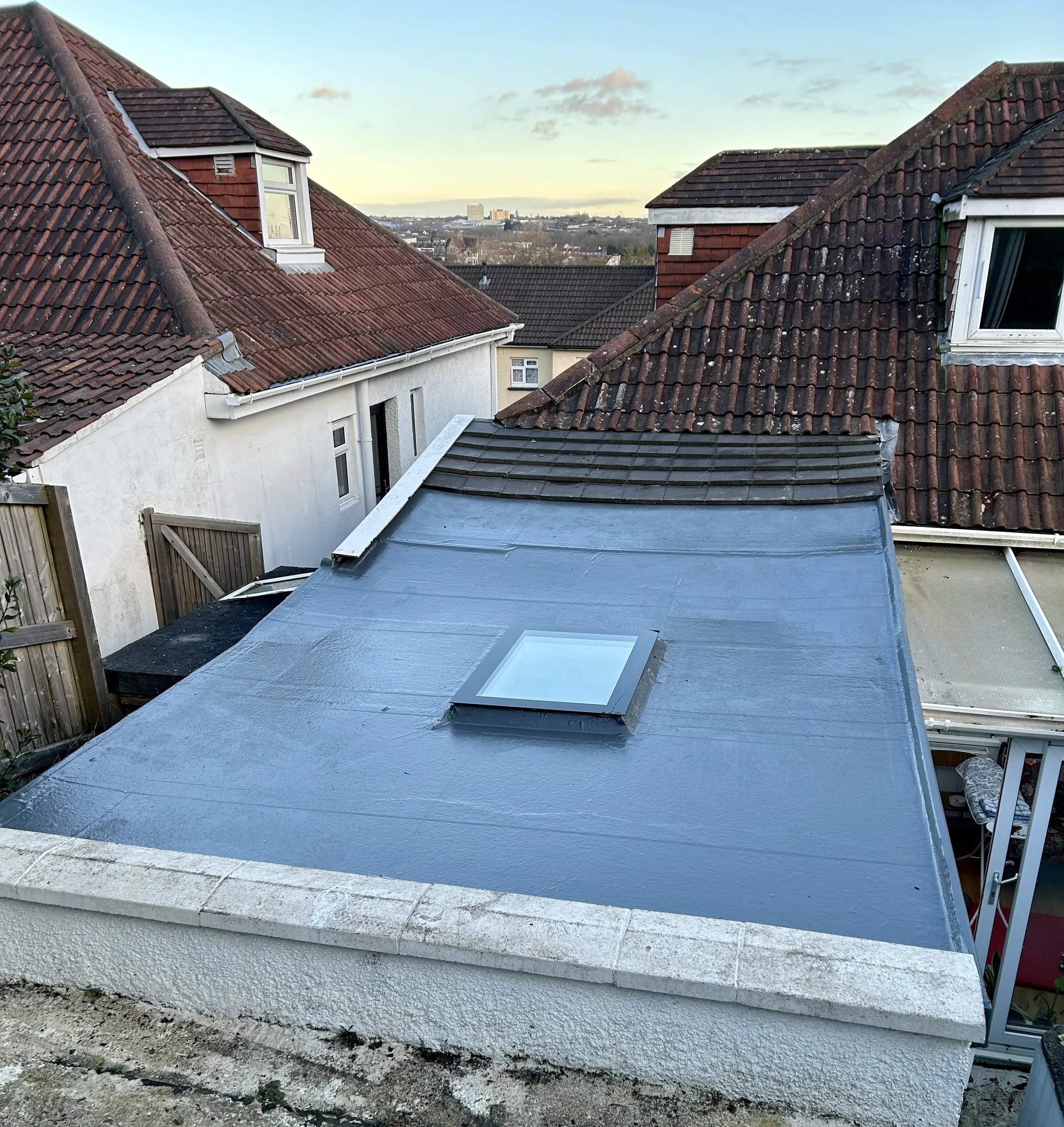 A view of a rooftop with dormer windows, some with open windows, in a residential area during daylight. There are other rooftops and city buildings in the distance, with a partly cloudy sky.