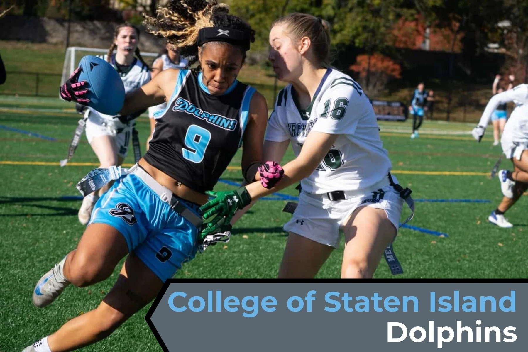 A female lacrosse player in a black and blue uniform, numbered 9, is running on a green field while holding a lacrosse stick. She is being pursued or guarded by another female player in a white uniform, numbered 16. In the background, there are other