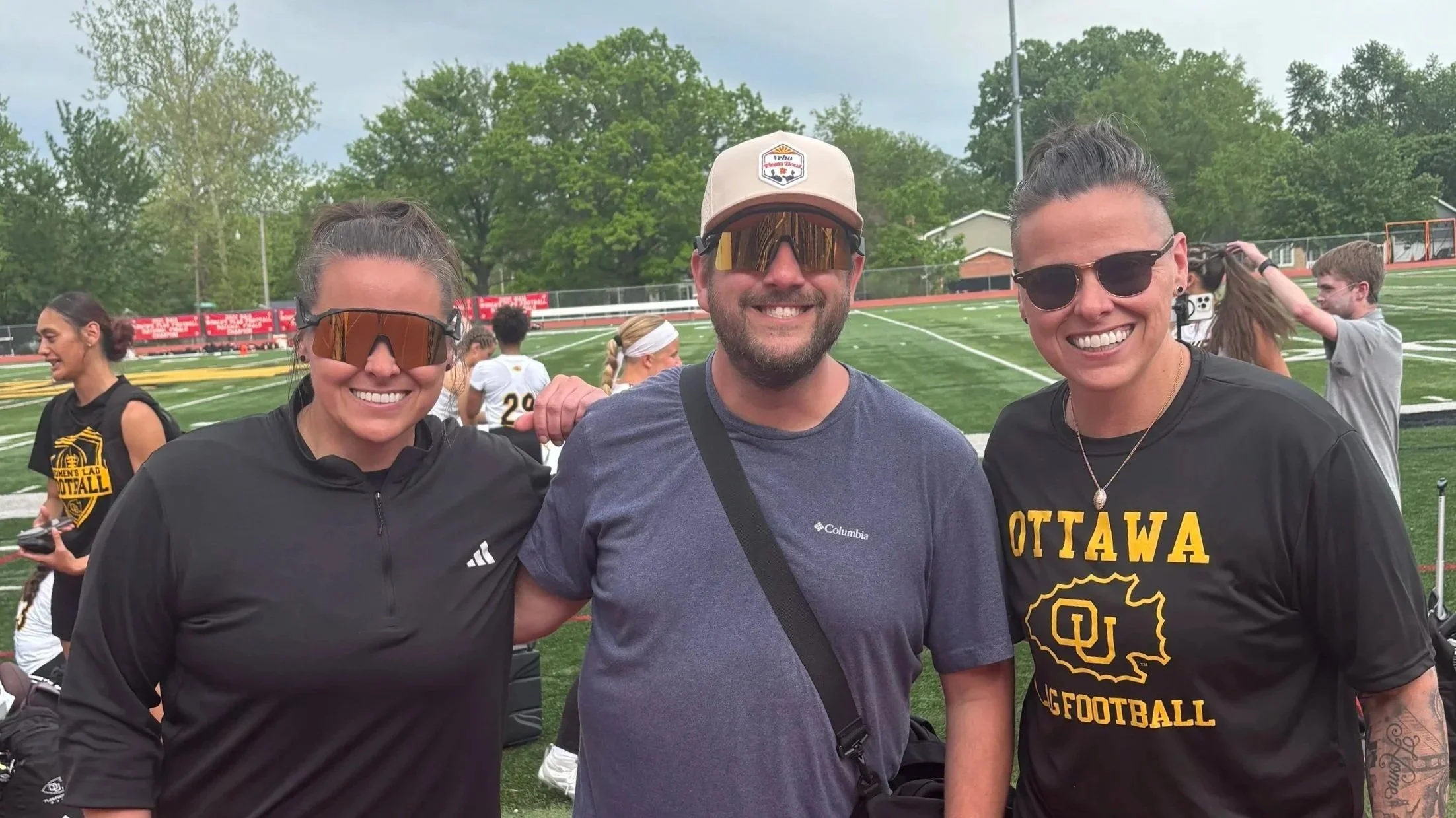 Three smiling people with sunglasses at a sports field, two women and one man, all wearing athletic clothing, with others in the background and trees around the field.