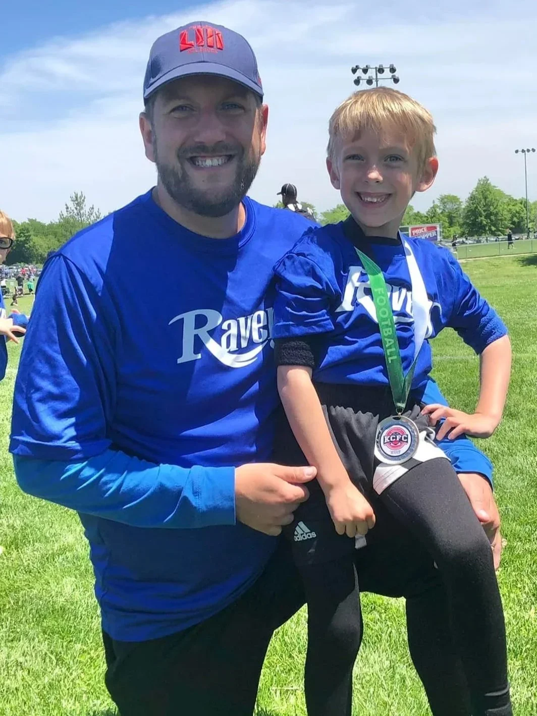 A man and a young boy at a sports field smiling, with the boy wearing a medal and a blue sports jersey. The man is wearing a blue shirt and a gray cap with red writing.