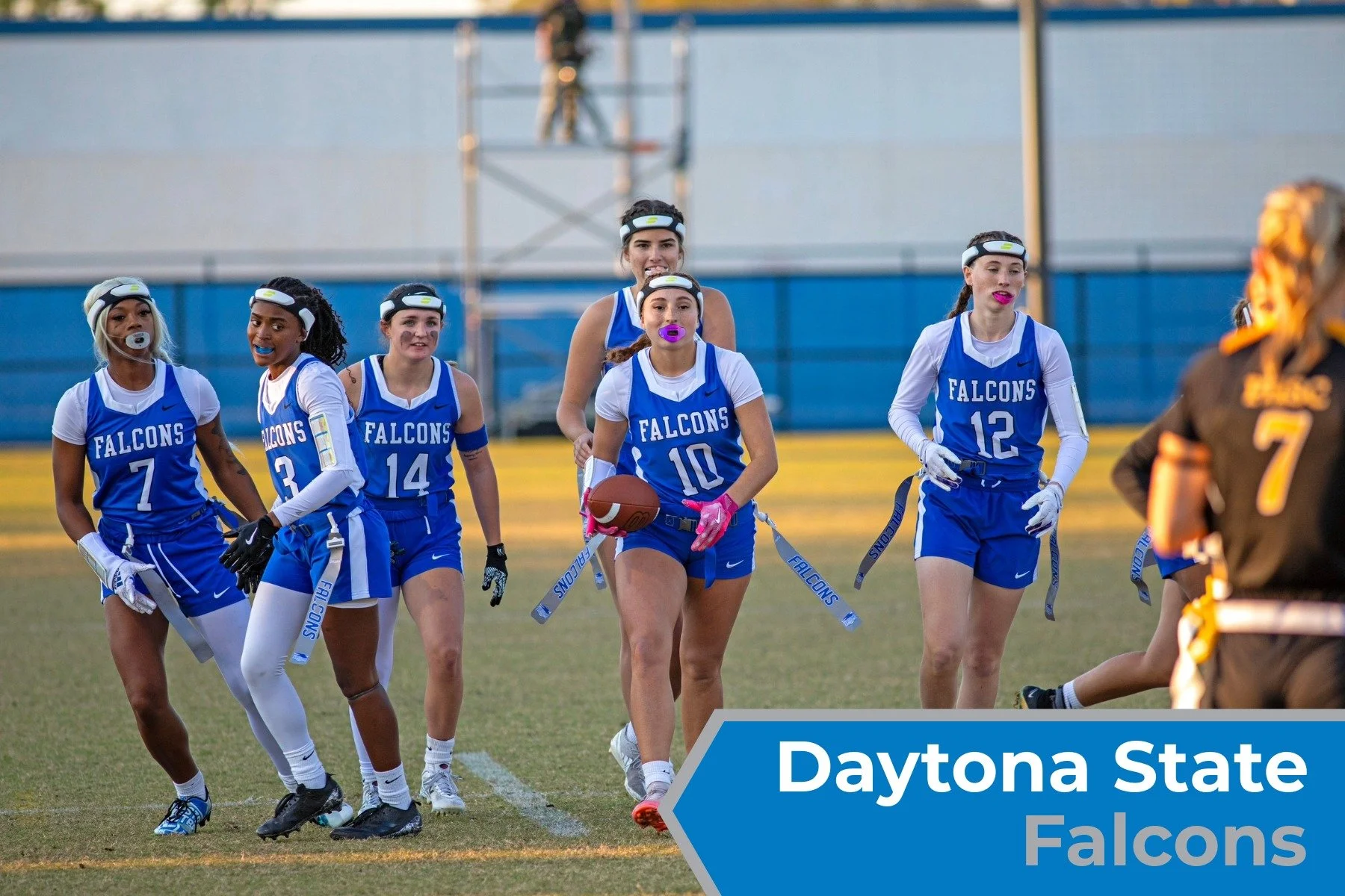 Women football players in blue jerseys with 'FALCONS' on them, walking on a field, with one holding a football and wearing pink gloves, during a game or practice, with a blue banner labeled 'Daytona State Falcons' in the foreground.