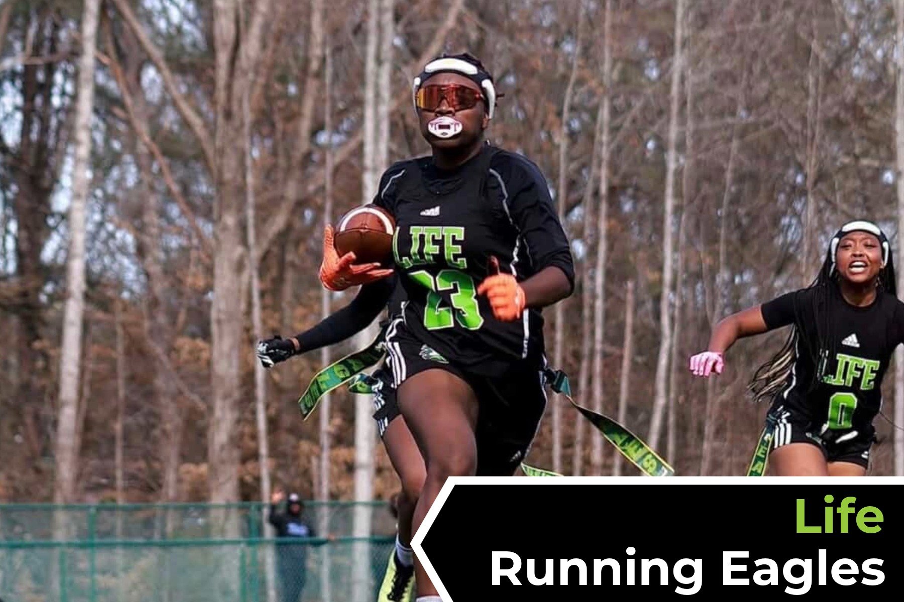 Young women running outdoors, one holding a football, wearing black sports jerseys with green lettering, in a wooded area.