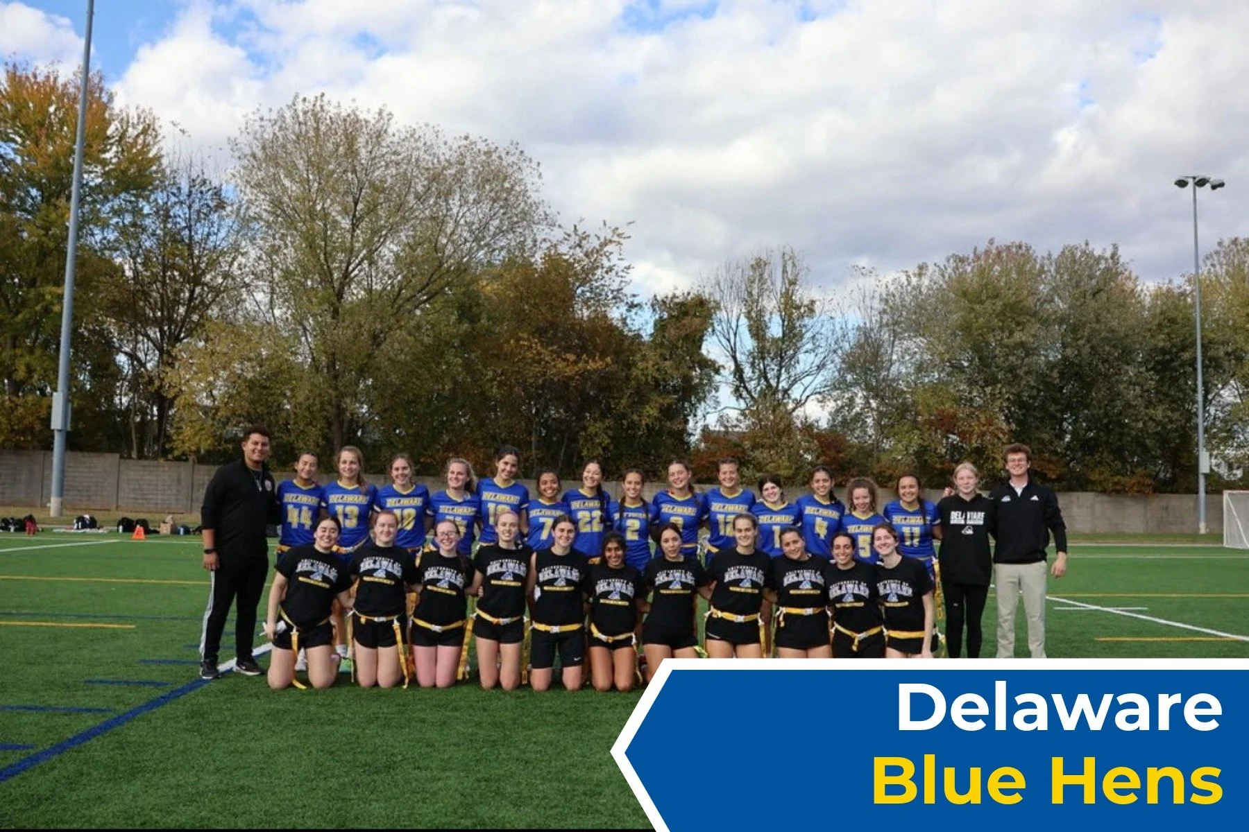 Group photo of a women's lacrosse team called Delaware Blue Hens, with players in blue and black uniforms, and two coaches standing on a football field with trees and cloudy sky in the background.