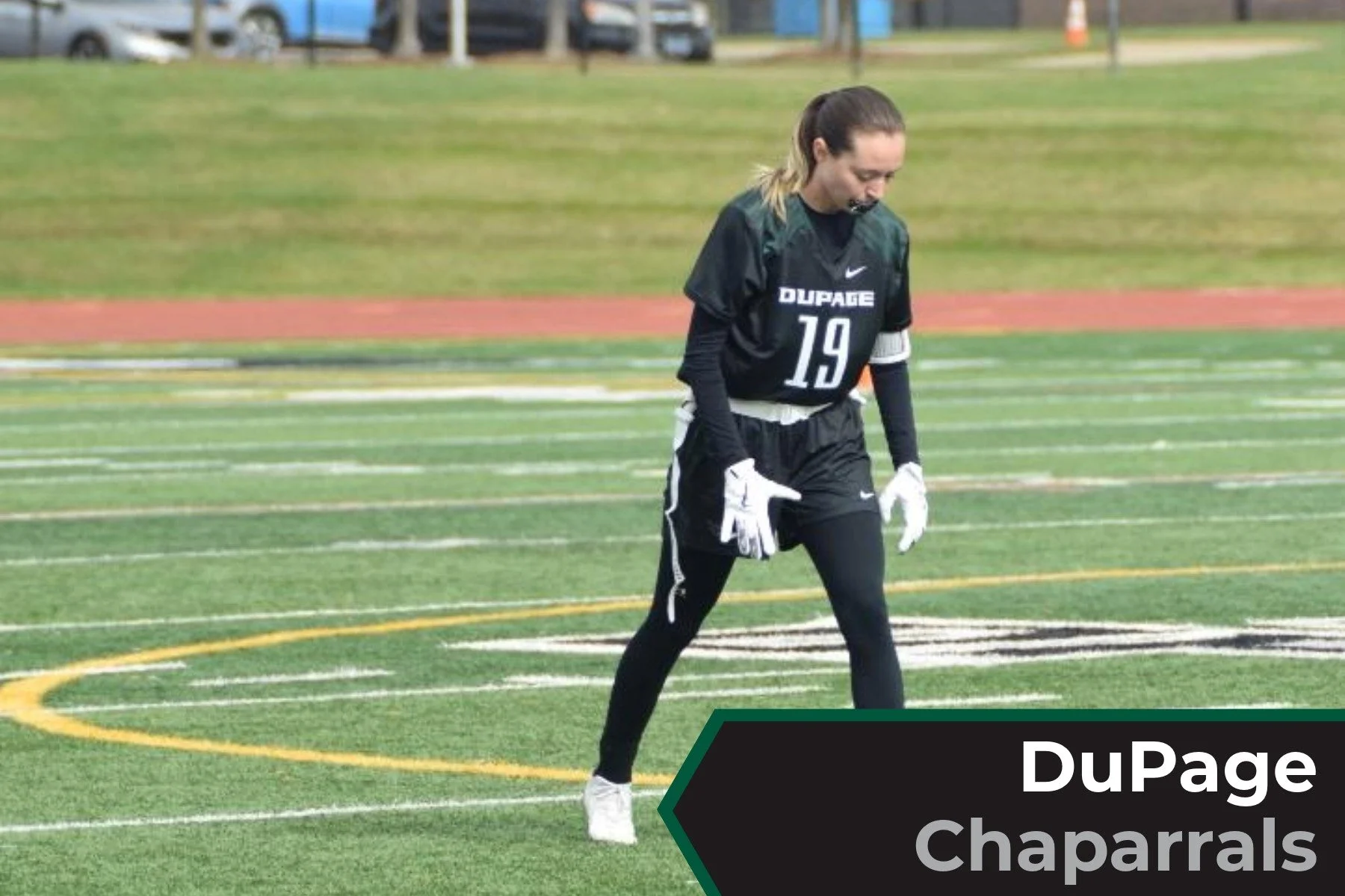 A female football player standing on a sports field wearing a black DuPage chaparrals jersey with the number 19, white gloves, and black leggings.