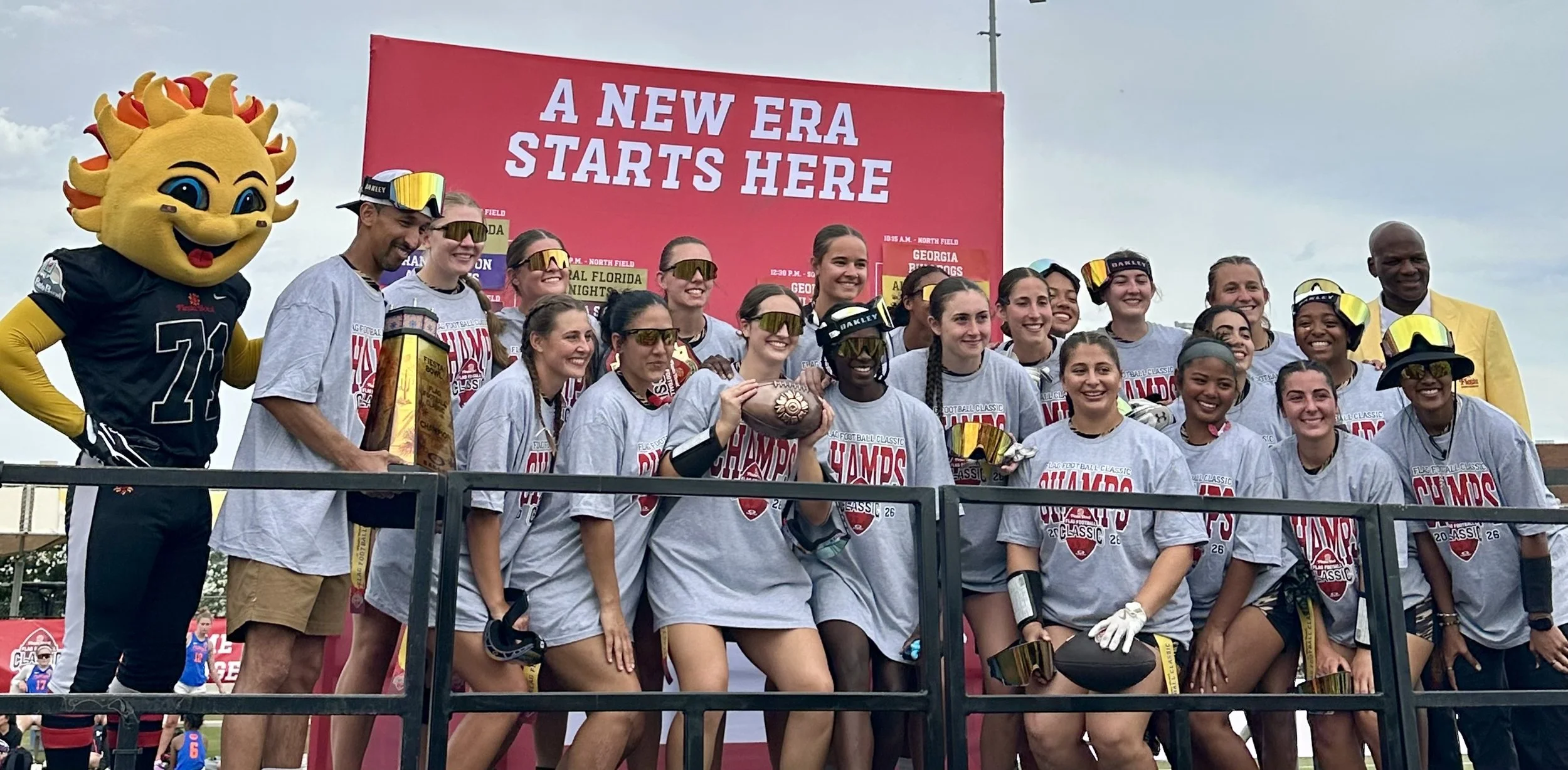 Group of female football players celebrating on a podium with trophies, wearing gray T-shirts and sunglasses, alongside a mascot with a sun face, and a man in a yellow suit, in front of a red sign that says 'A NEW ERA STARTS HERE'.