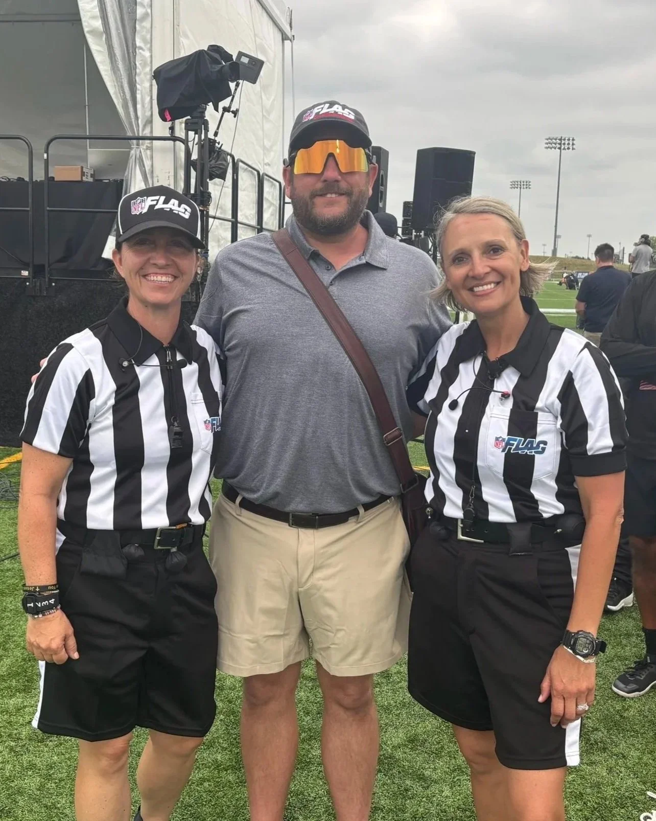 Three people standing on a sports field: two women dressed as referees in black and white striped shirts, and a man in casual clothing, all smiling and posing for the photo.