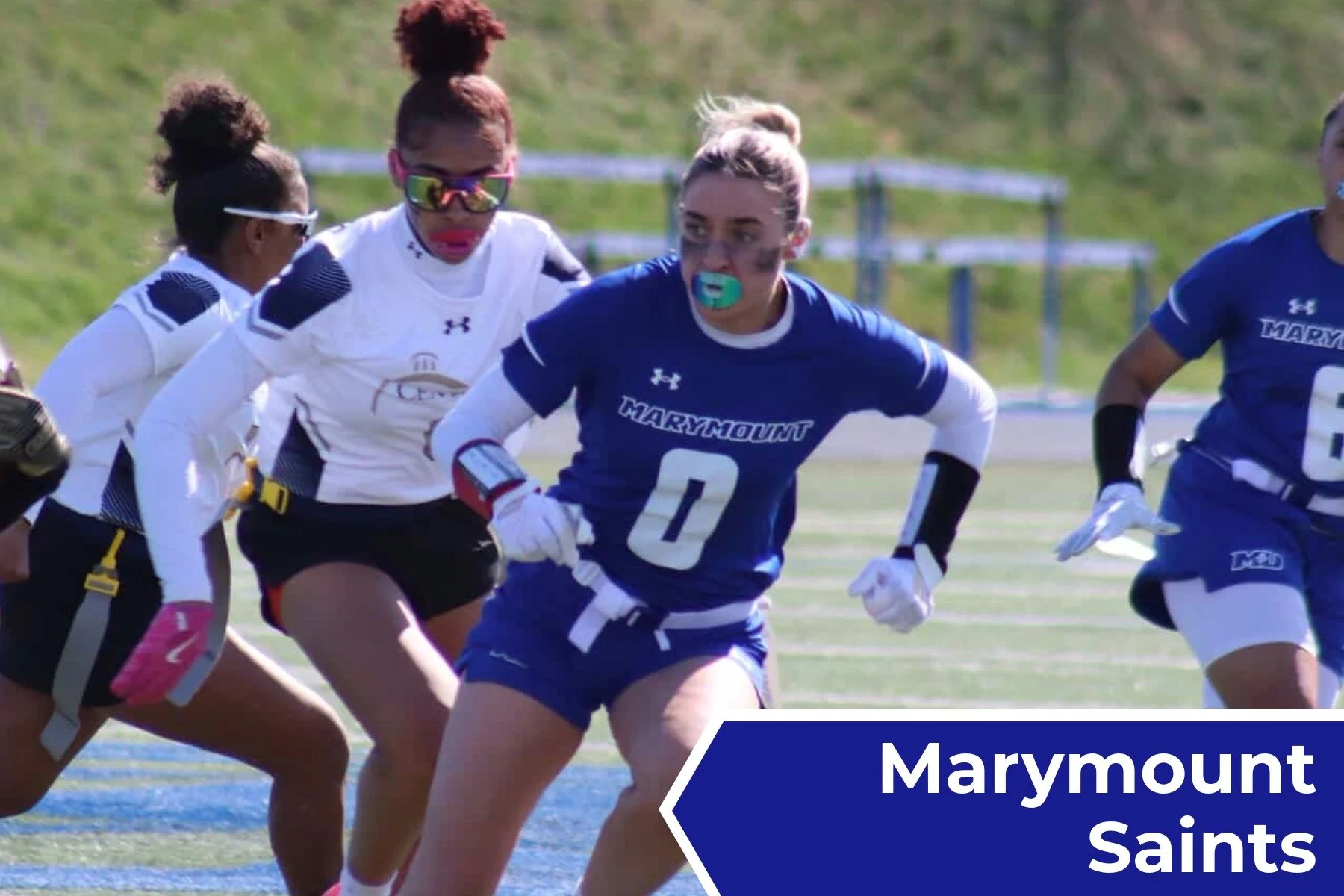 Women football players in uniform and gloves, running on a football field during a game, with a blue and white banner reading 'Marymount Saints'.
