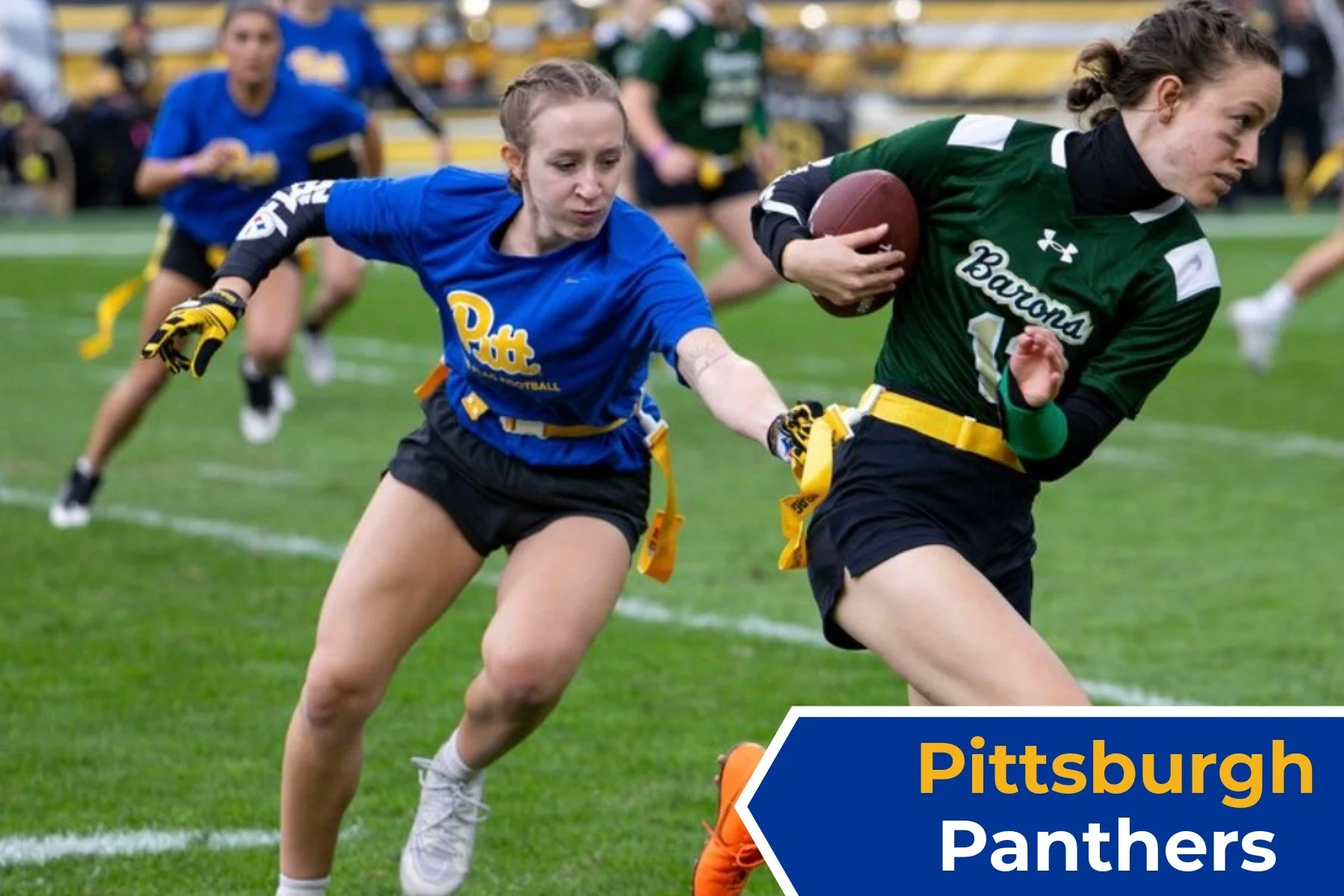 A female football player in a green uniform with 'Barons' written on it is running with a football, while another female player in a blue uniform with 'Pitt' is reaching out to grab the football from her on a football field. Other players are visible