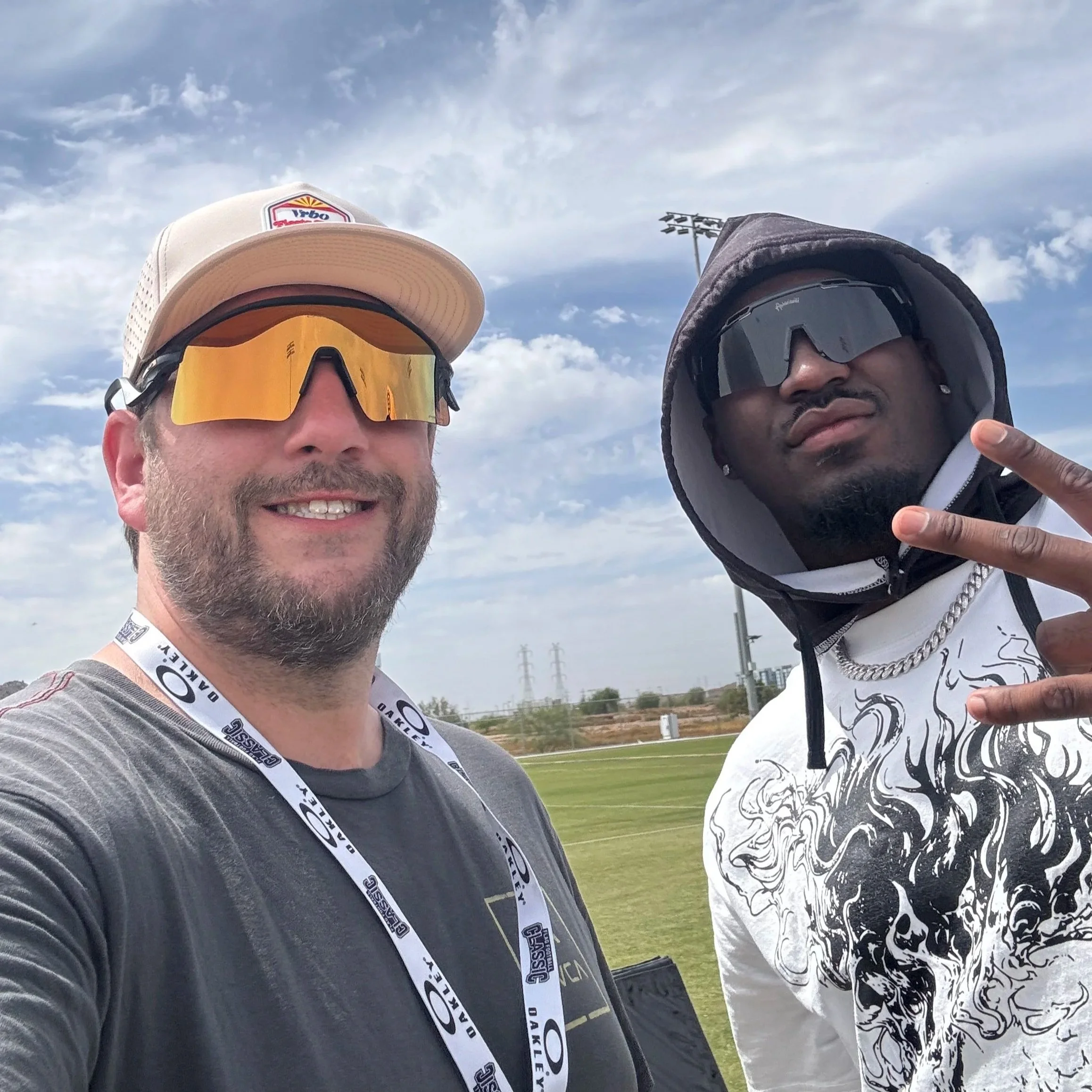 Two men wearing sunglasses and caps posing for a selfie outdoors, with a cloudy sky and stadium lights in the background.