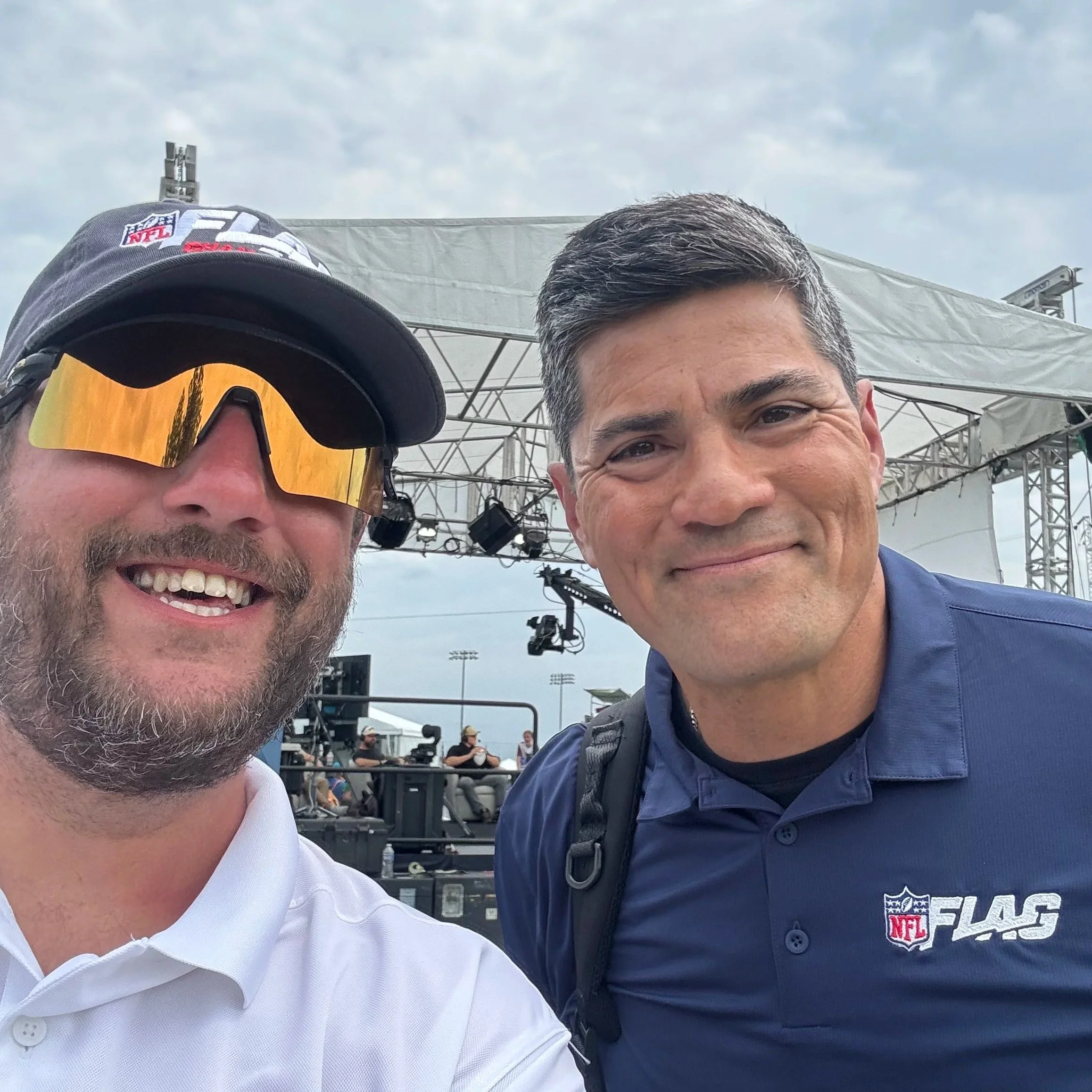 Close-up of two men smiling at a sports event, one wearing a white shirt and the other wearing a navy NFL jersey, with a stage and cloudy sky in the background.