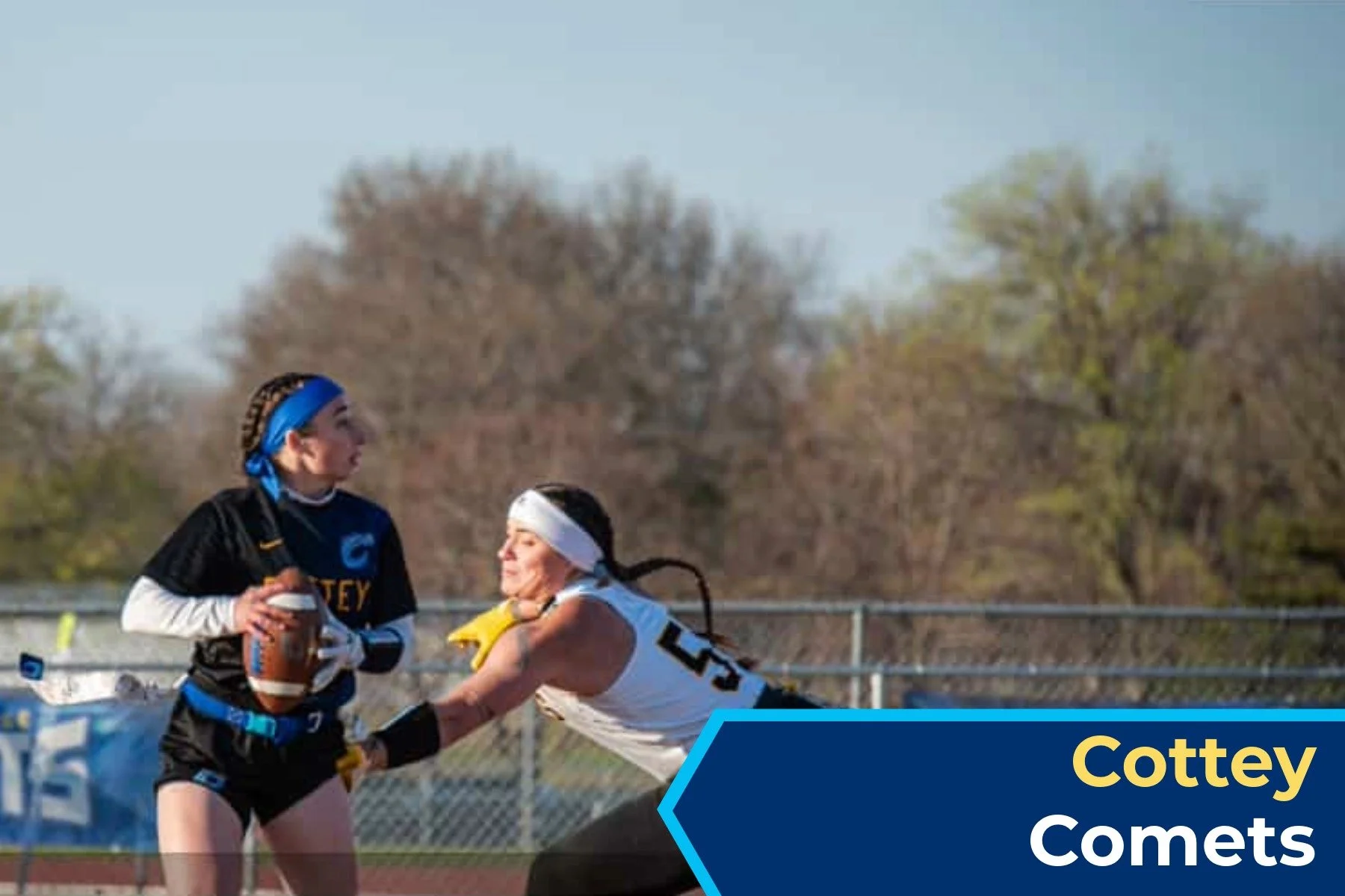 Two female athletes playing flag football on a field, with trees in the background, one in a black and blue uniform and the other in a white uniform wearing a headband. The player in black is holding a football, while the player in white is reaching 