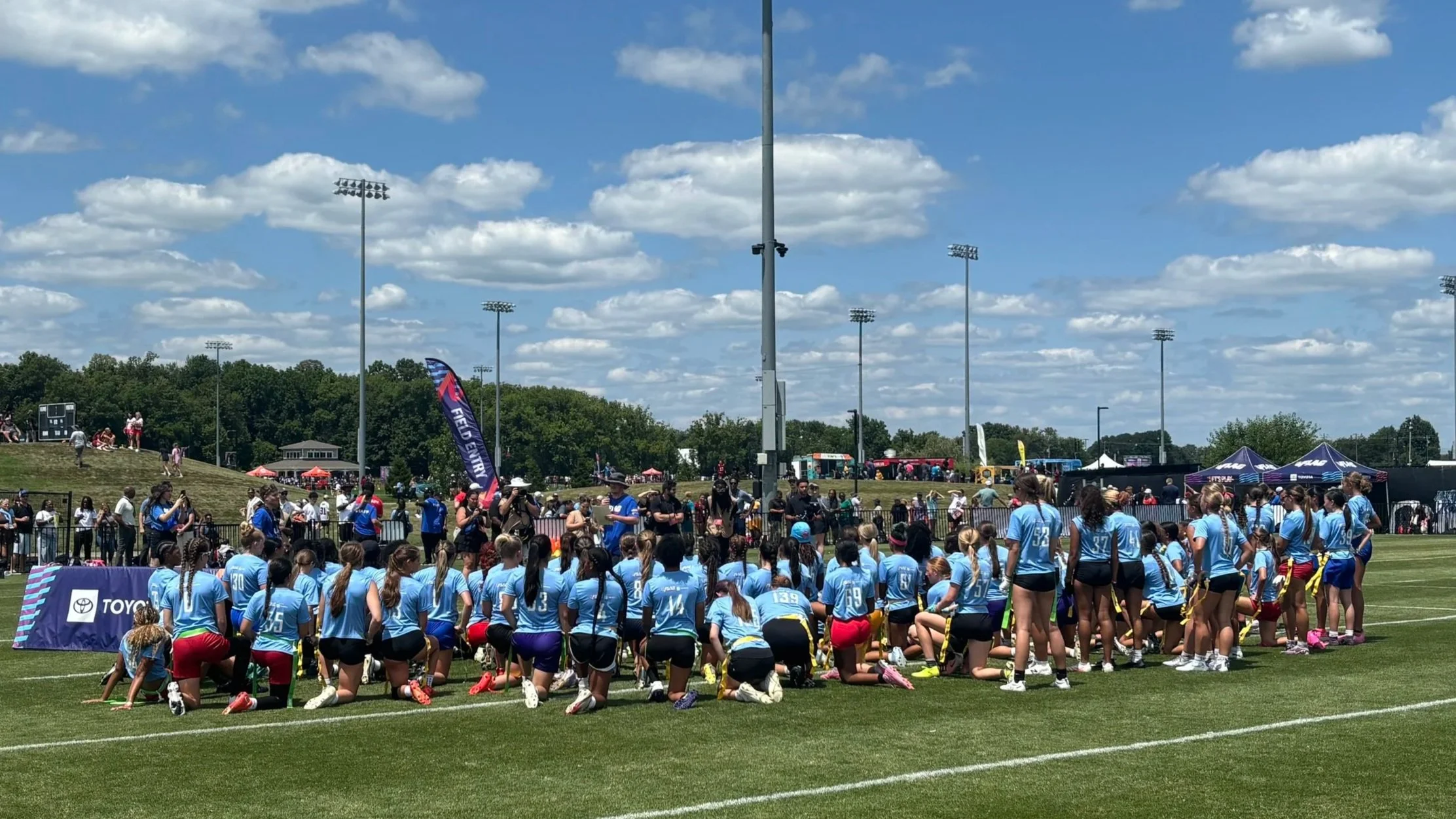 A large group of young female athletes wearing blue sports uniforms gathered on a grassy field at an outdoor sports event on a sunny day with a partly cloudy sky. Spectators and other participants are visible in the background, along with tents, banners, and tall floodlights.