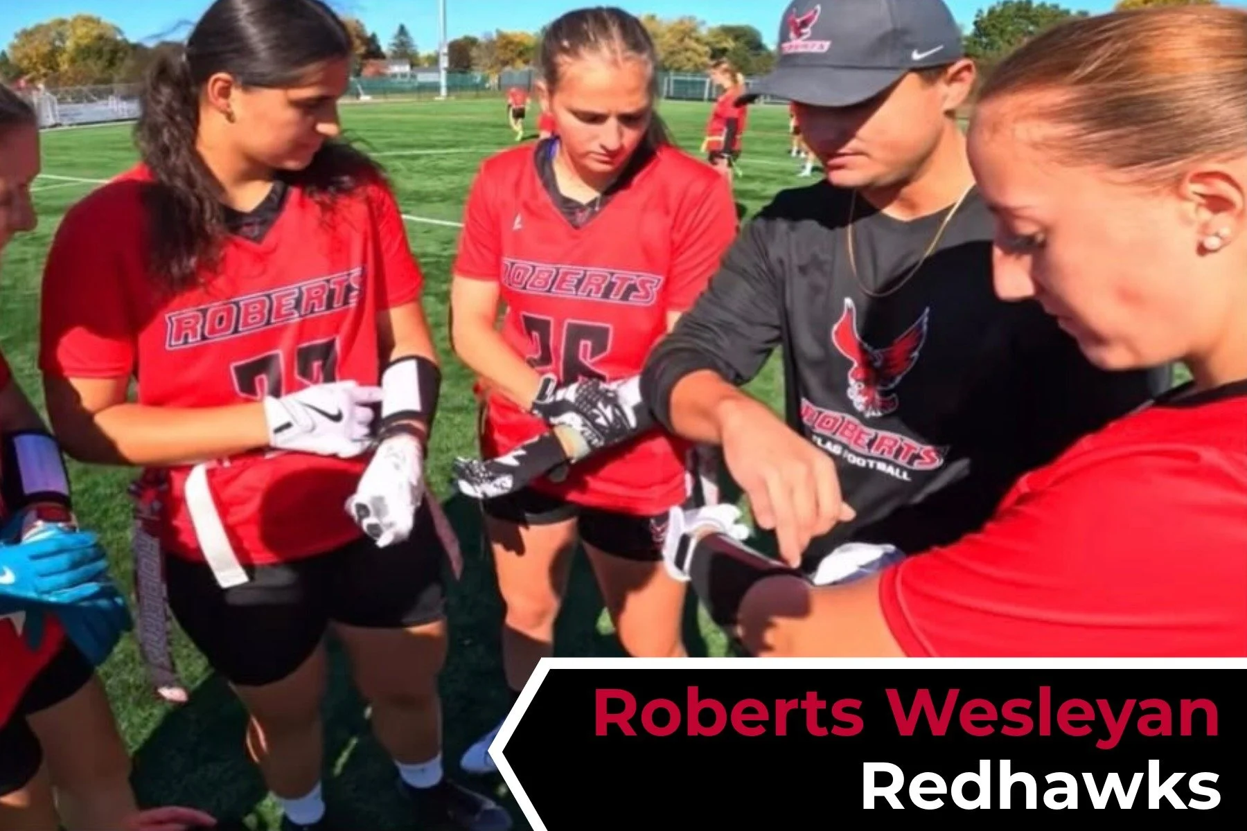 A group of female football players in red jerseys with 'Roberts' and numbers, and their coach, on a grassy football field during daytime. They are engaged in a discussion, with some wearing gloves and one holding a football helmet.
