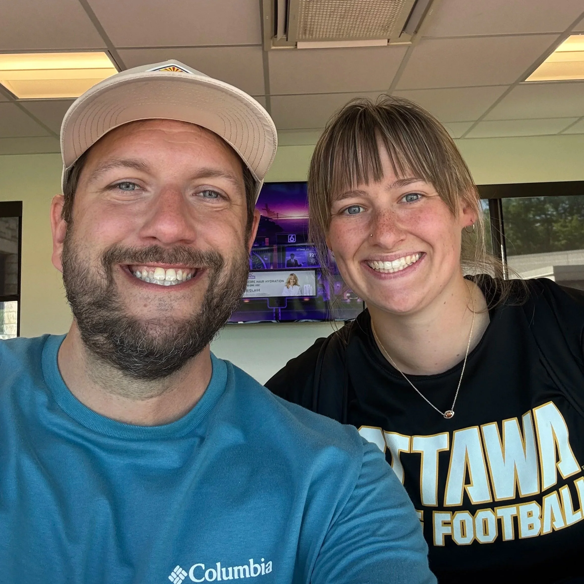 Close-up of a man and a woman smiling, with the man wearing a beige cap and a blue Columbia shirt, and the woman with short brown hair, freckles, and a black shirt with yellow lettering, in an indoor setting with a ceiling and a television displaying a purple sky and a building in the background.