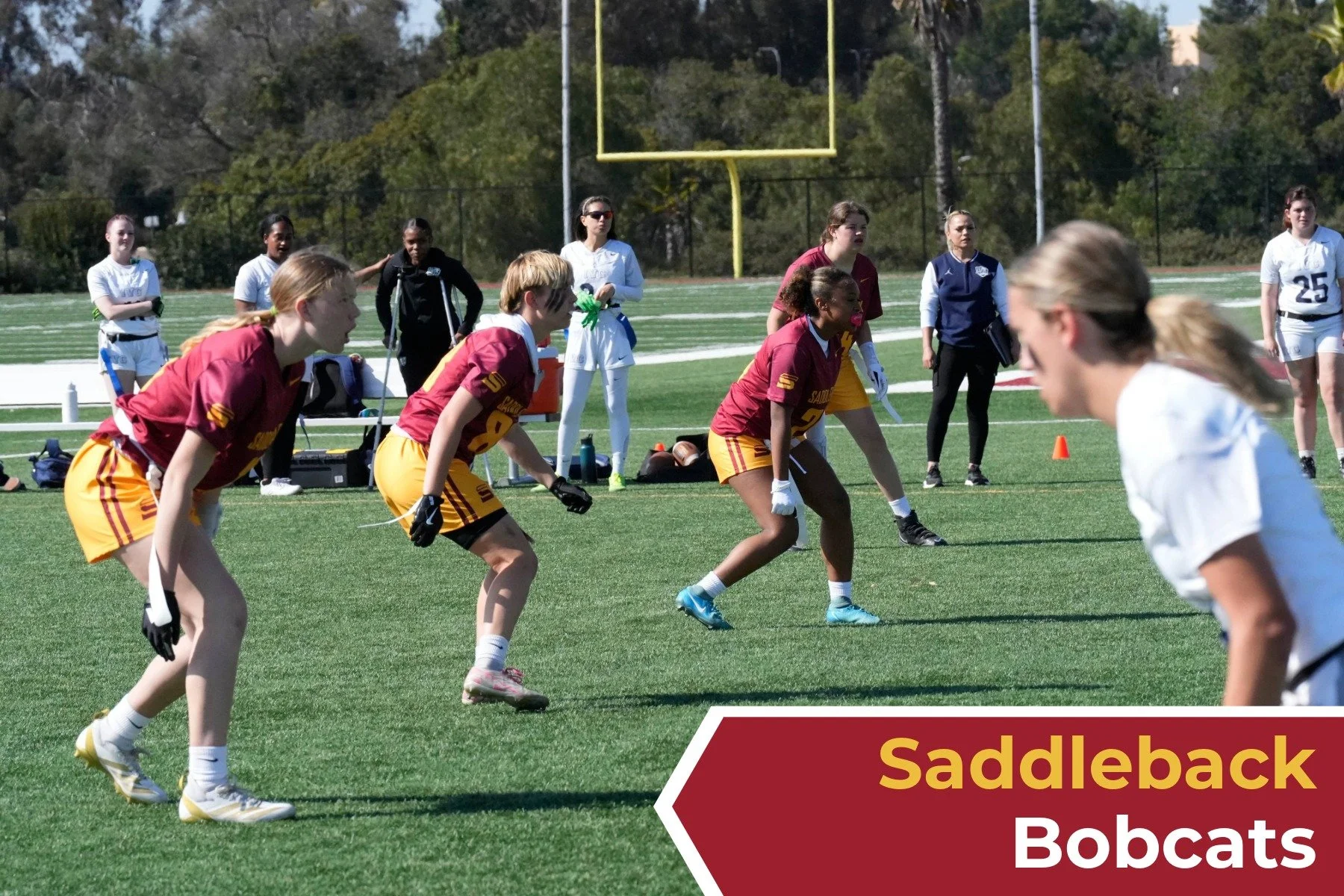 Women playing football on a field with a group of spectators and greenery in the background. The image features players in maroon and white uniforms, with a focus on a woman in white about to race across the field. The overlay text reads 'Saddleback 
