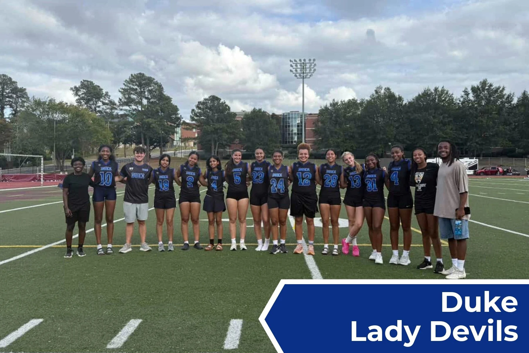 A group of female volleyball players in black and blue uniforms standing on a football field, with a coach or team manager at the right end, during daytime under a cloudy sky. There is a graphic label in the bottom right corner that says 'Duke Lady D