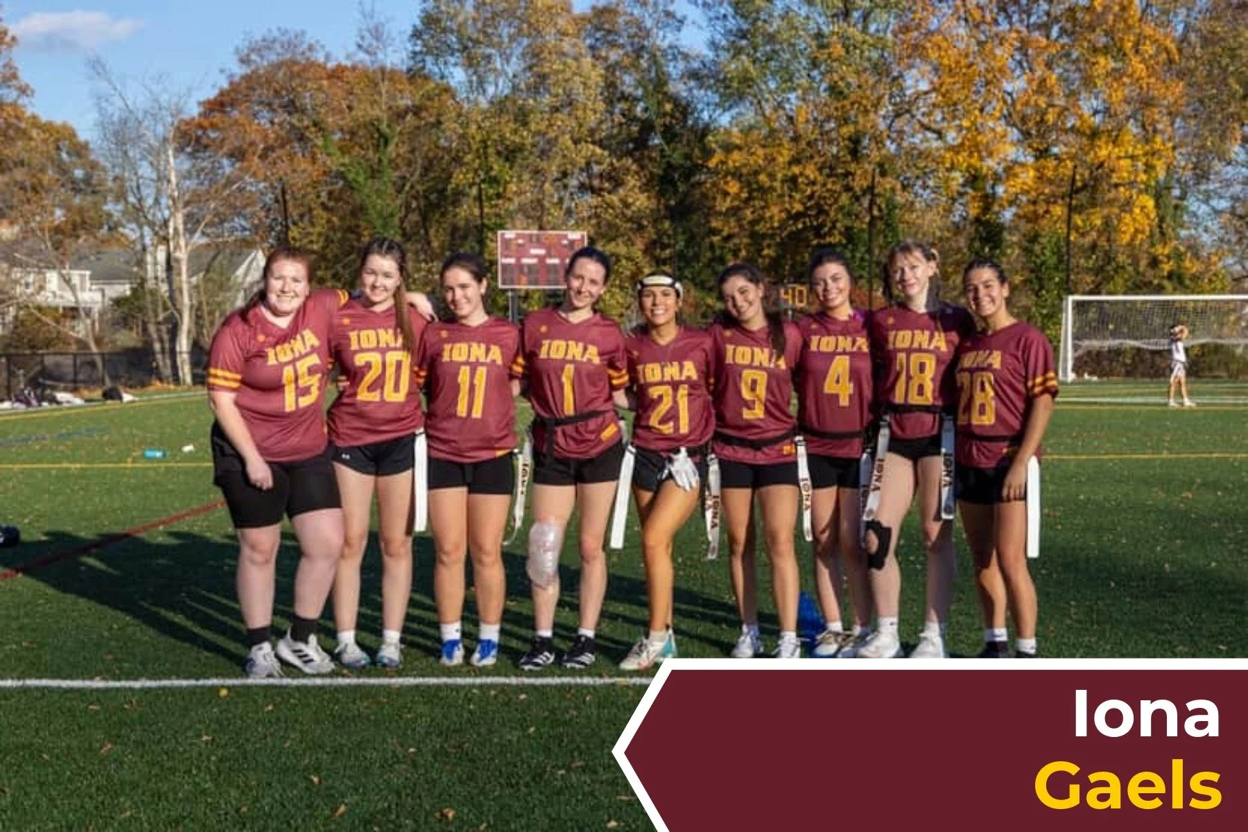 A team of Womens College flag football players for the Iona Gaels in maroon and yellow uniforms standing on a field, smiling with arms around each other, with fall foliage in the background.