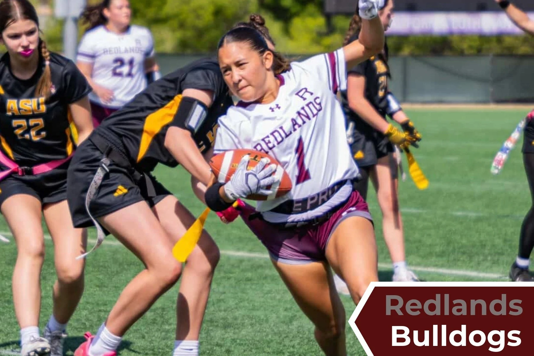 A woman in a white and maroon football jersey with 'Redlands' and the number 1 on it, holding a football and running on a grassy field during a women's football game.