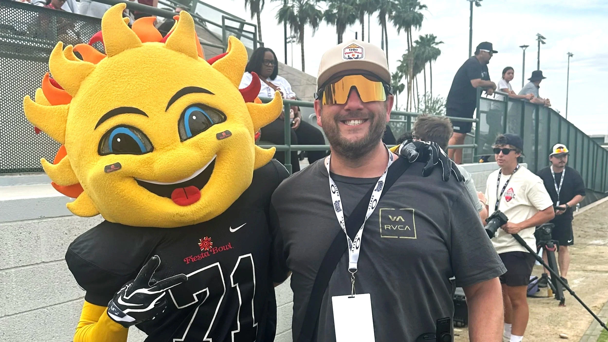 A man smiling while standing next to a person in a sun mascot costume at a sports event. The mascot has a large yellow face with sun-shaped rays, large blue eyes, and a friendly smile. Background shows spectators and palm trees, indicating an outdoor setting.