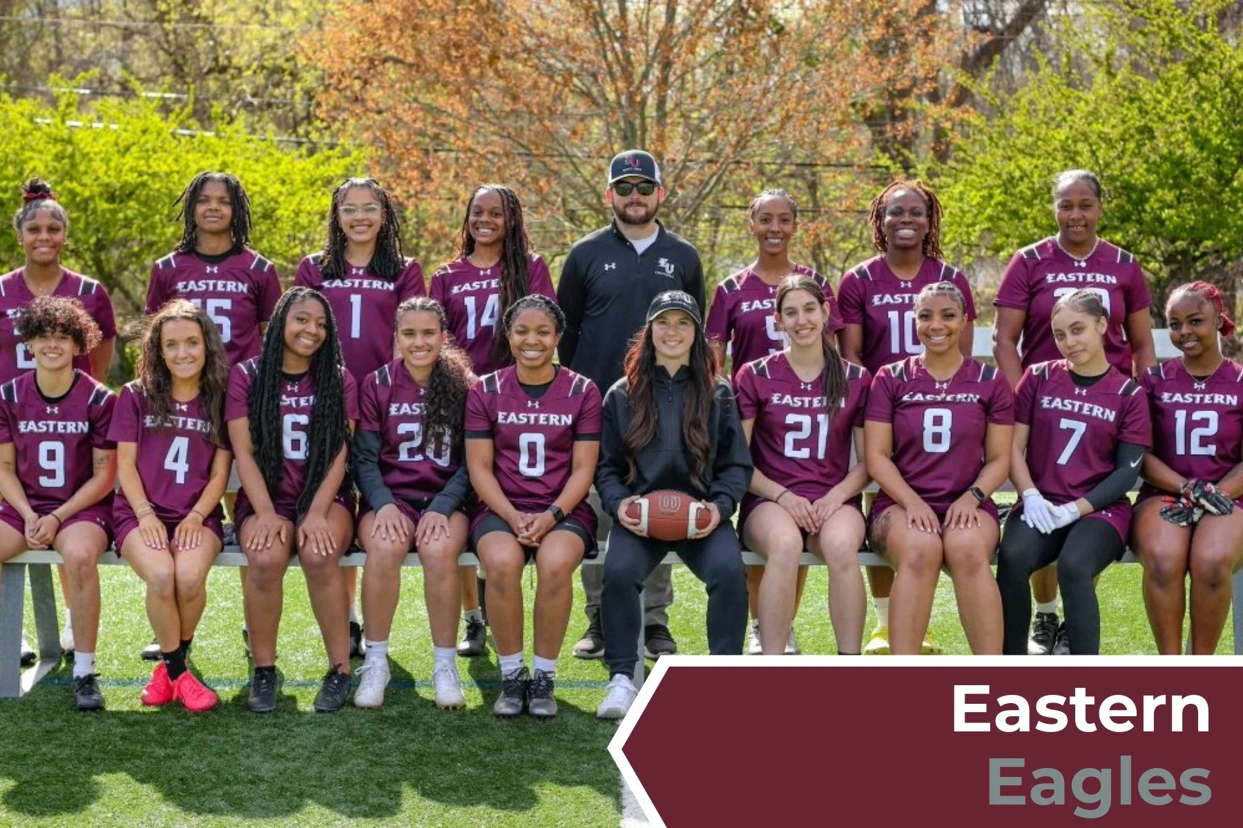 Group photo of the Eastern Eagles women's football team outdoors with trees in the background, wearing maroon uniforms and some holding a football.