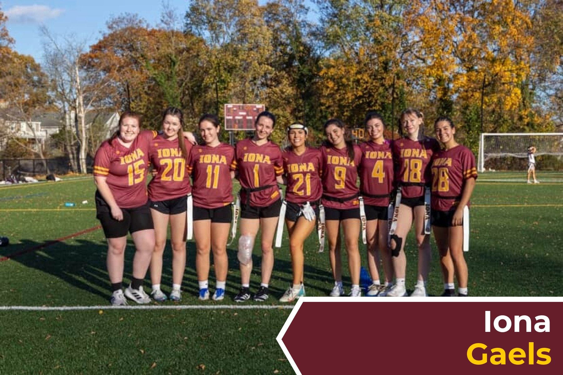 A group of nine female soccer players standing on a soccer field, wearing maroon and gold uniforms, with trees in fall colors in the background.