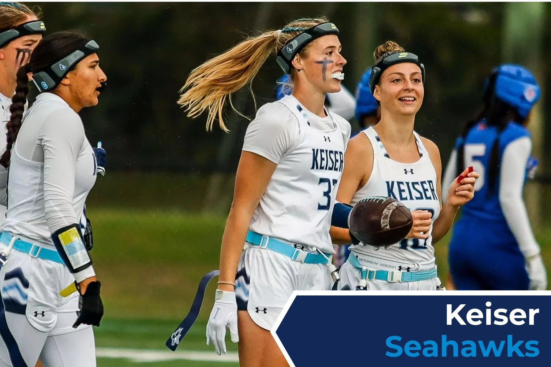 Image of young women playing American football, wearing white and blue uniforms with 'Keiser Seahawks' written on their jerseys, helmets, and gear, on a grassy field.