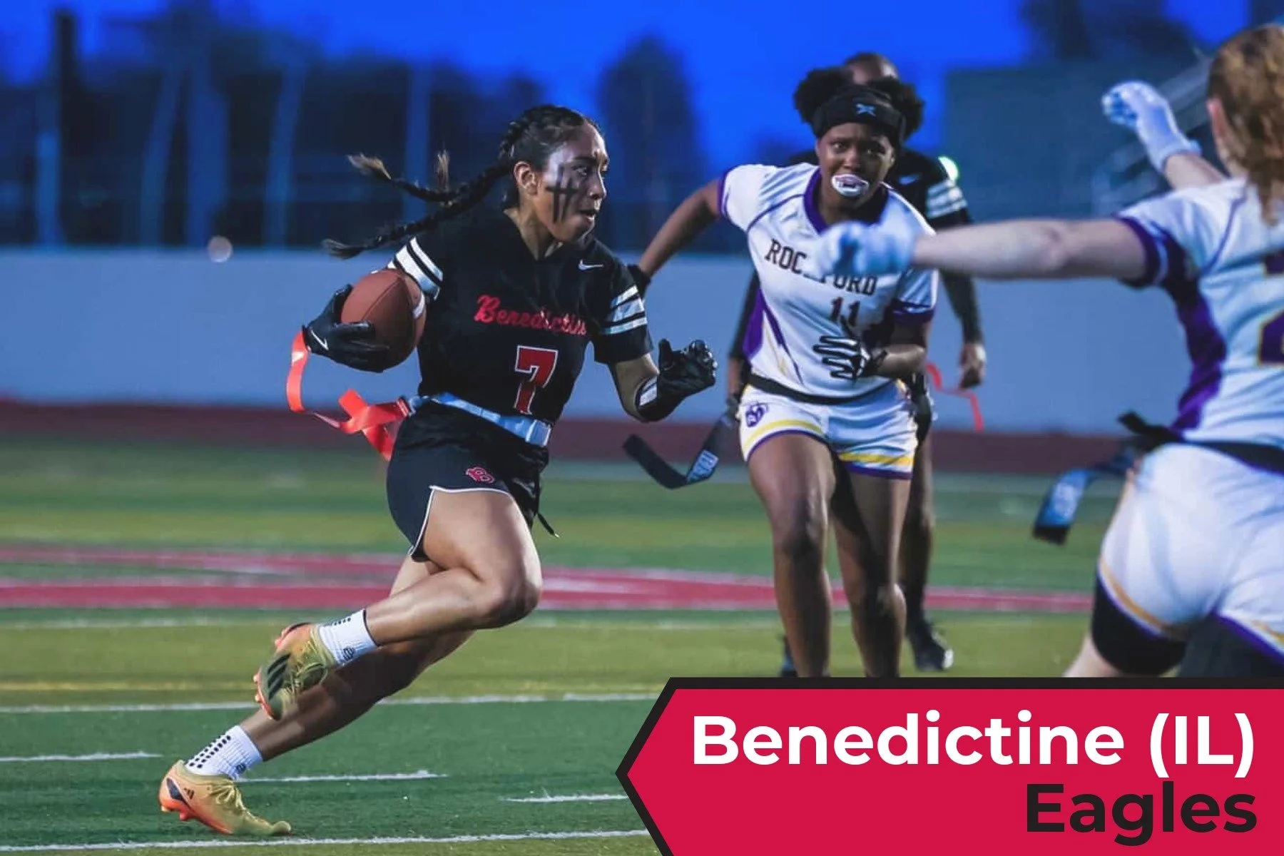 Girls playing flag football on a field during dusk. One girl is running with a ball, while others are trying to stop or block her. The girl with the ball has black face paint and is wearing a black uniform with red accents. The other girls are in whi