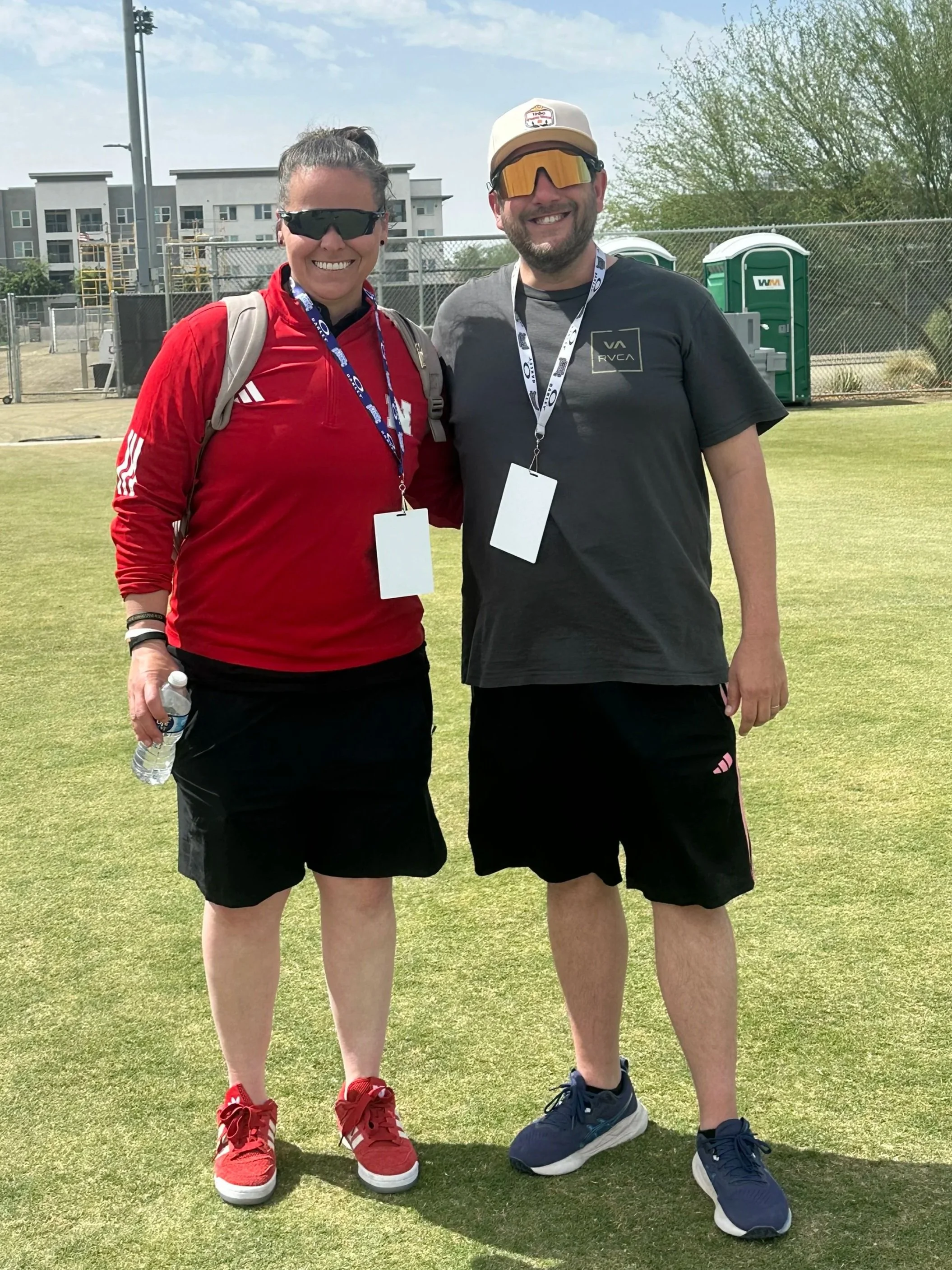 A woman and a man standing on a grass field, smiling and looking at the camera, wearing athletic clothing and sunglasses, with a background of a fence, trees, portable toilets, and a building.