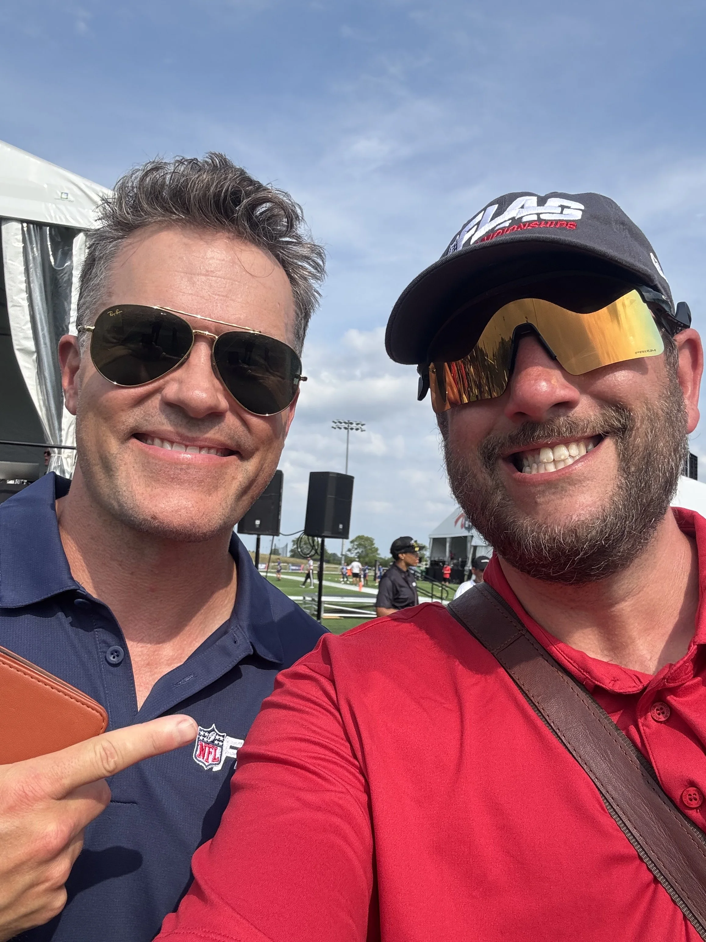 Two men smiling for a selfie, wearing sunglasses and NFL gear, in an outdoor setting with blue sky and clouds in the background, at what appears to be a stadium or event.