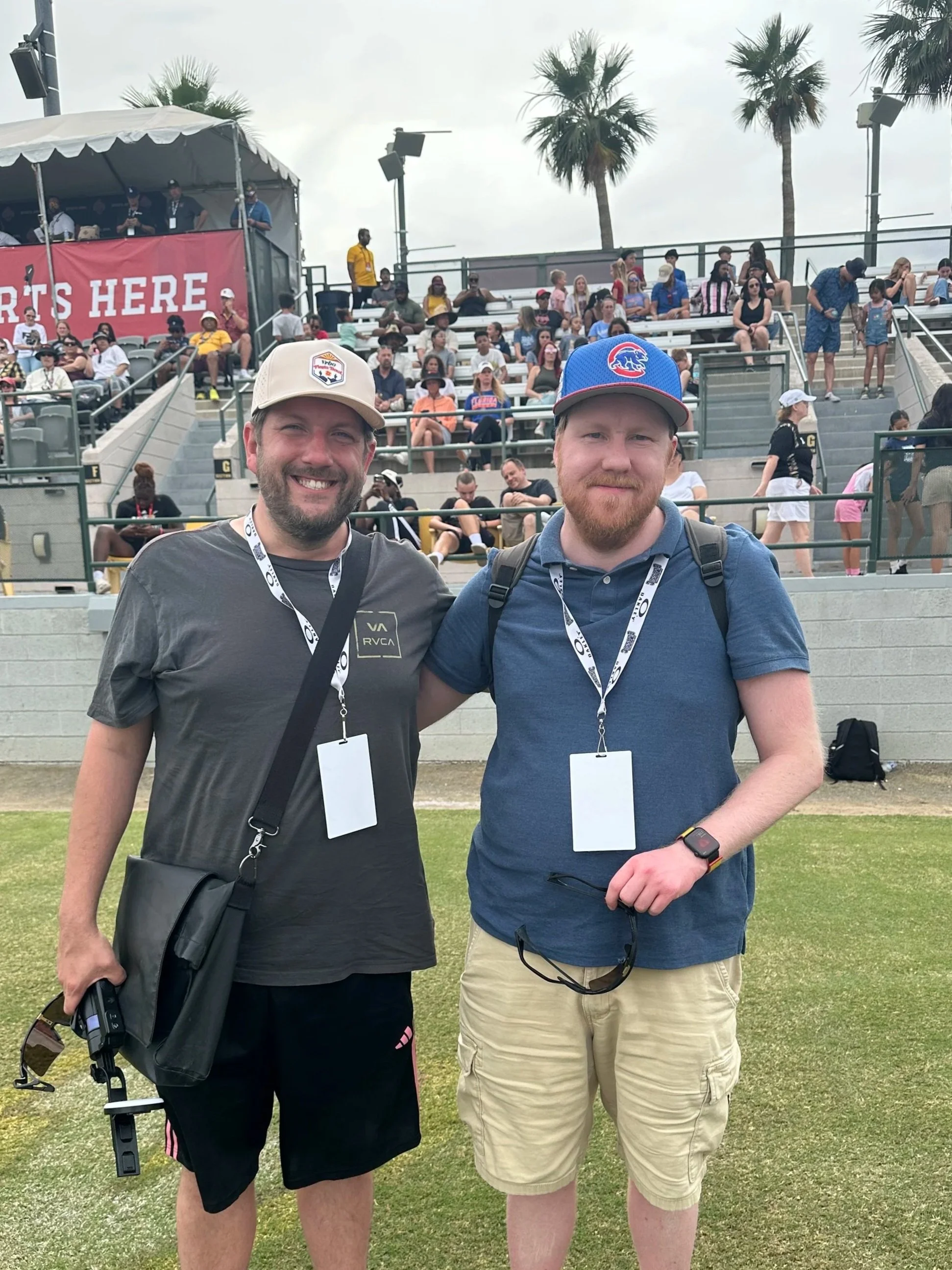 Two men standing on grass at a sports event, smiling at the camera, with many spectators in the bleachers behind them, and palm trees in the background.